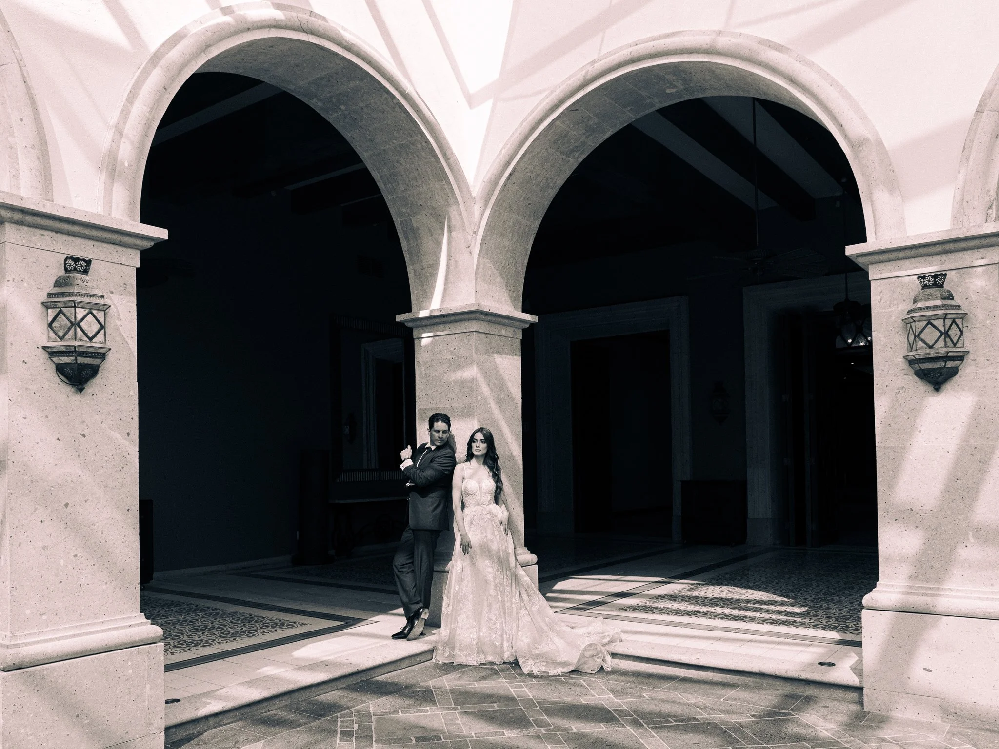 Pareja elegante, hombre con traje y mujer en vestido de novia, posando bajo arcos en un edificio sofisticado.