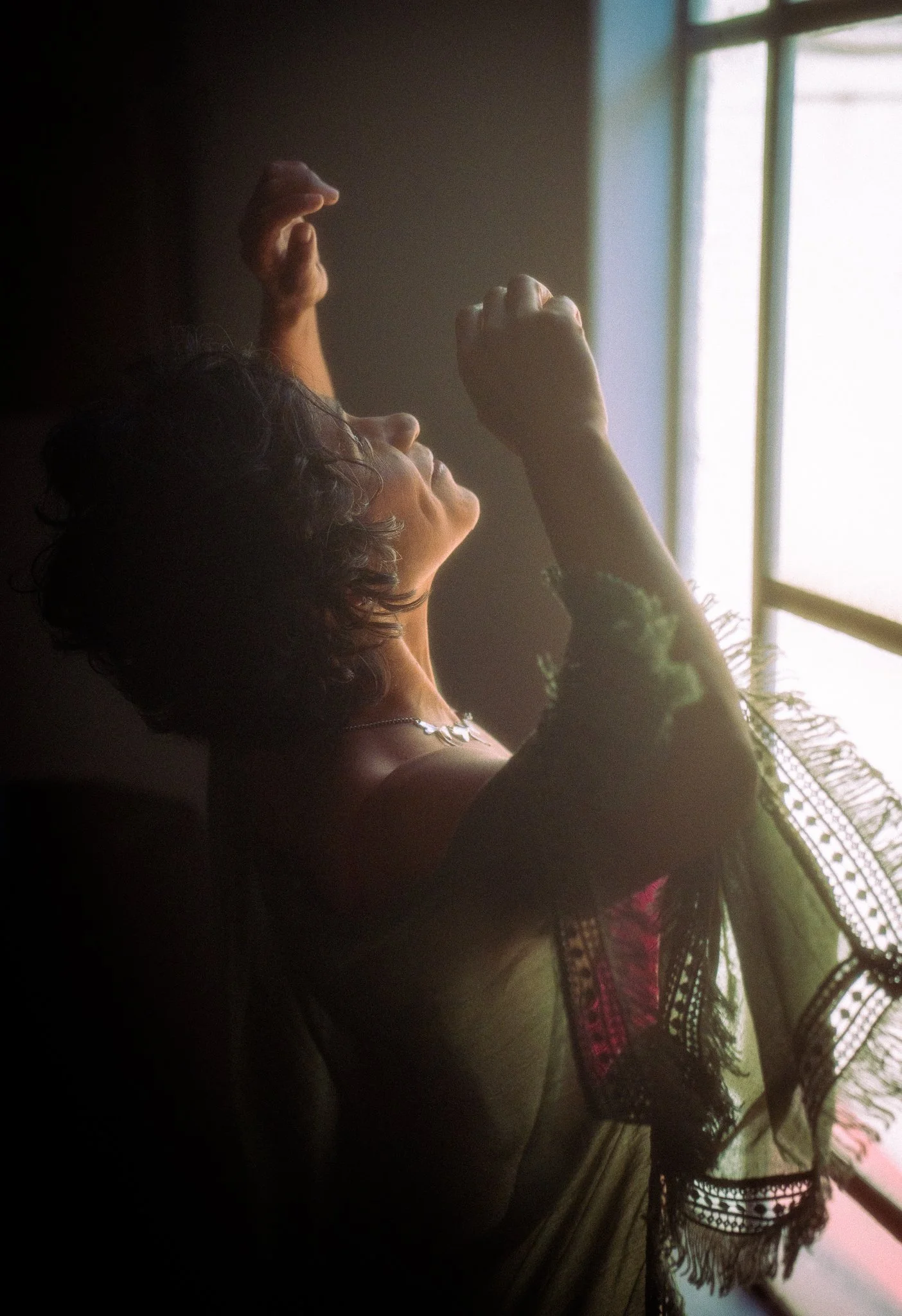 Mujer con cabello rizado, cerca de una ventana, iluminada por la luz natural, levantando las manos en una pose expresiva.