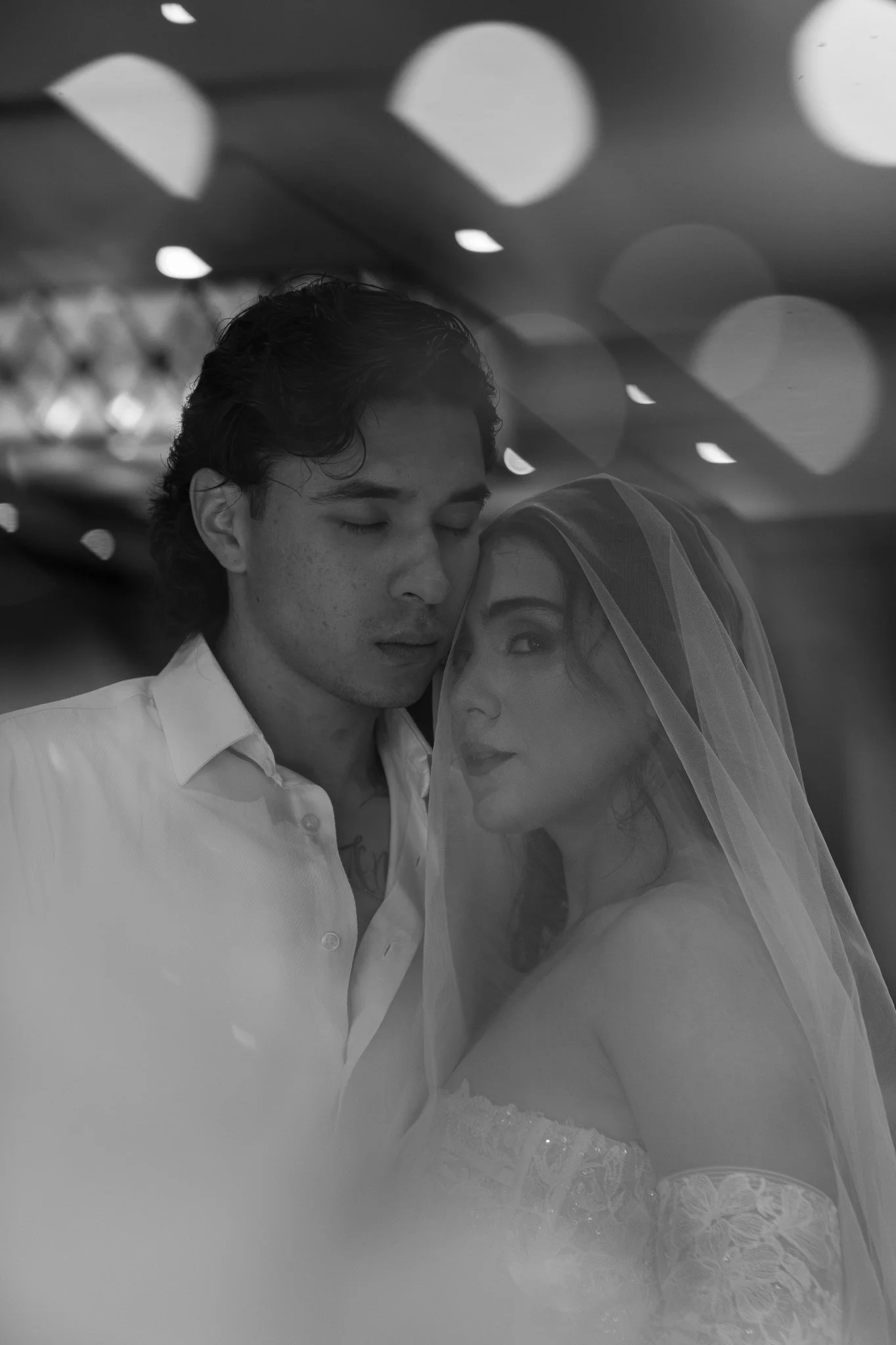 Una pareja en una boda, con el hombre con los ojos cerrados y la mujer con un velo, en un ambiente con luces suaves y borrosas.