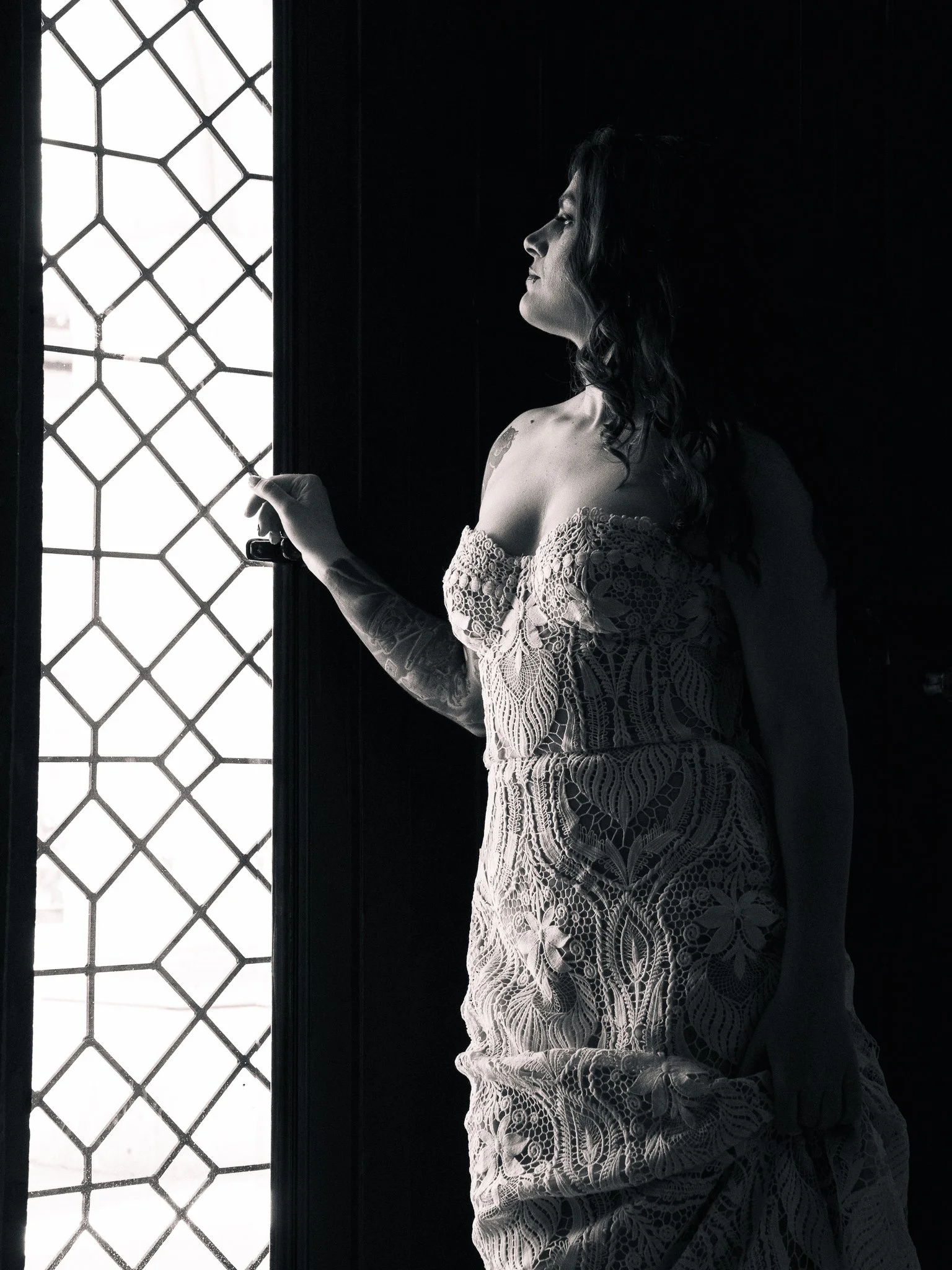 Mujer con vestido de encaje, de perfil, mirando por una ventana con estructura de rejas, en un ambiente oscuro