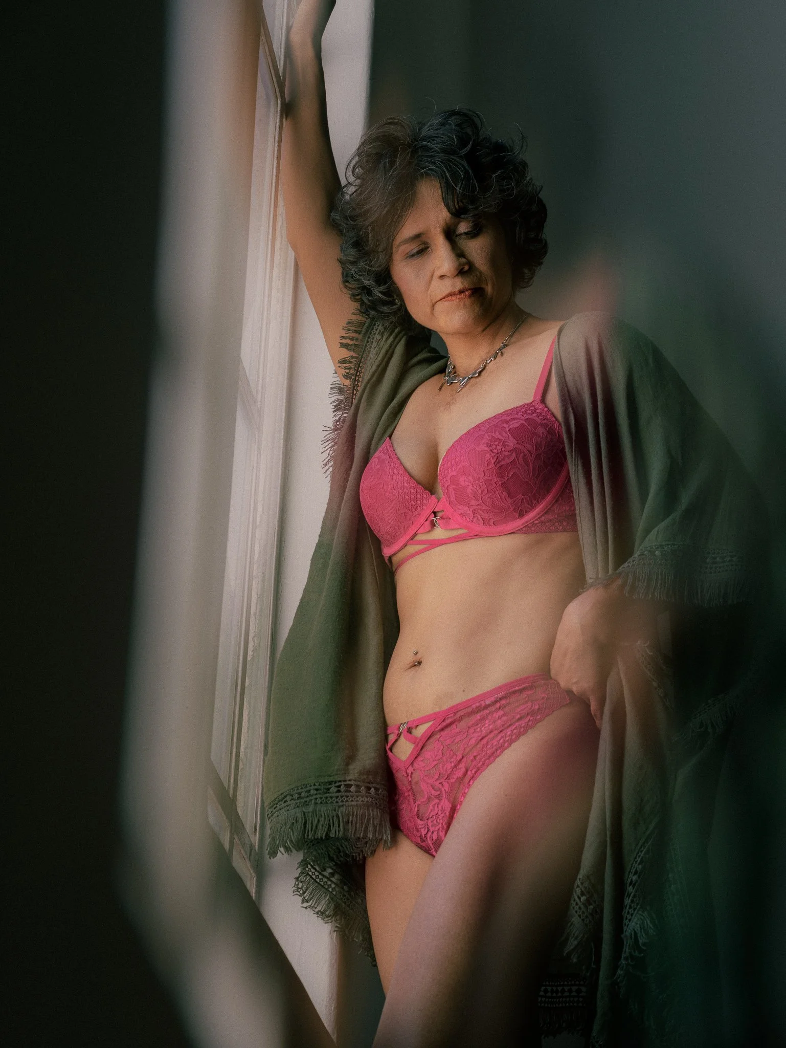 Mujer joven con ropa interior rosa y un abrigo verde, recargada contra una ventana, con expresión pensativa.
