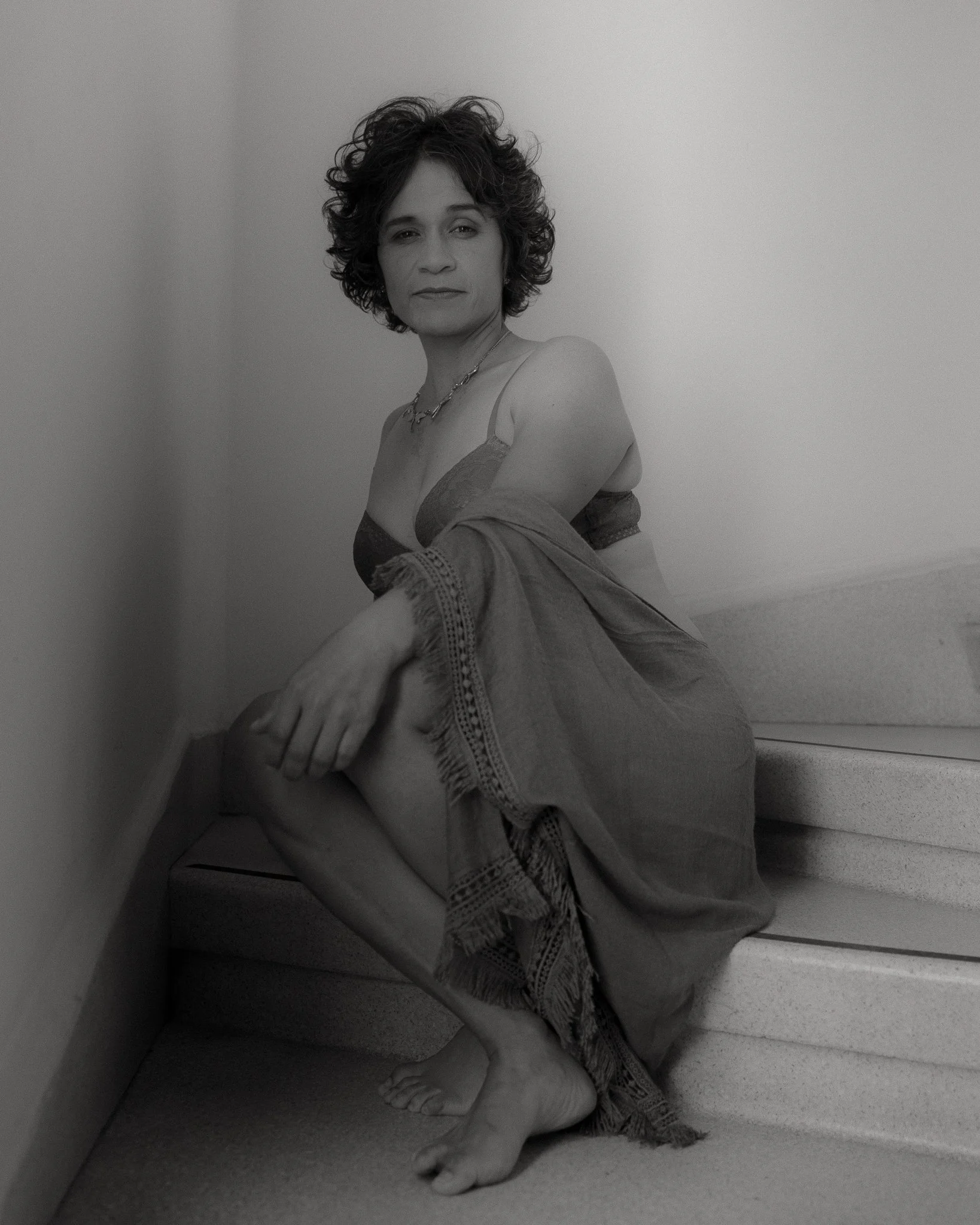 Mujer con cabello rizado oscuro, vestido y collar, sentada en las escaleras, en una pose contemplativa en la fotografía en blanco y negro.
