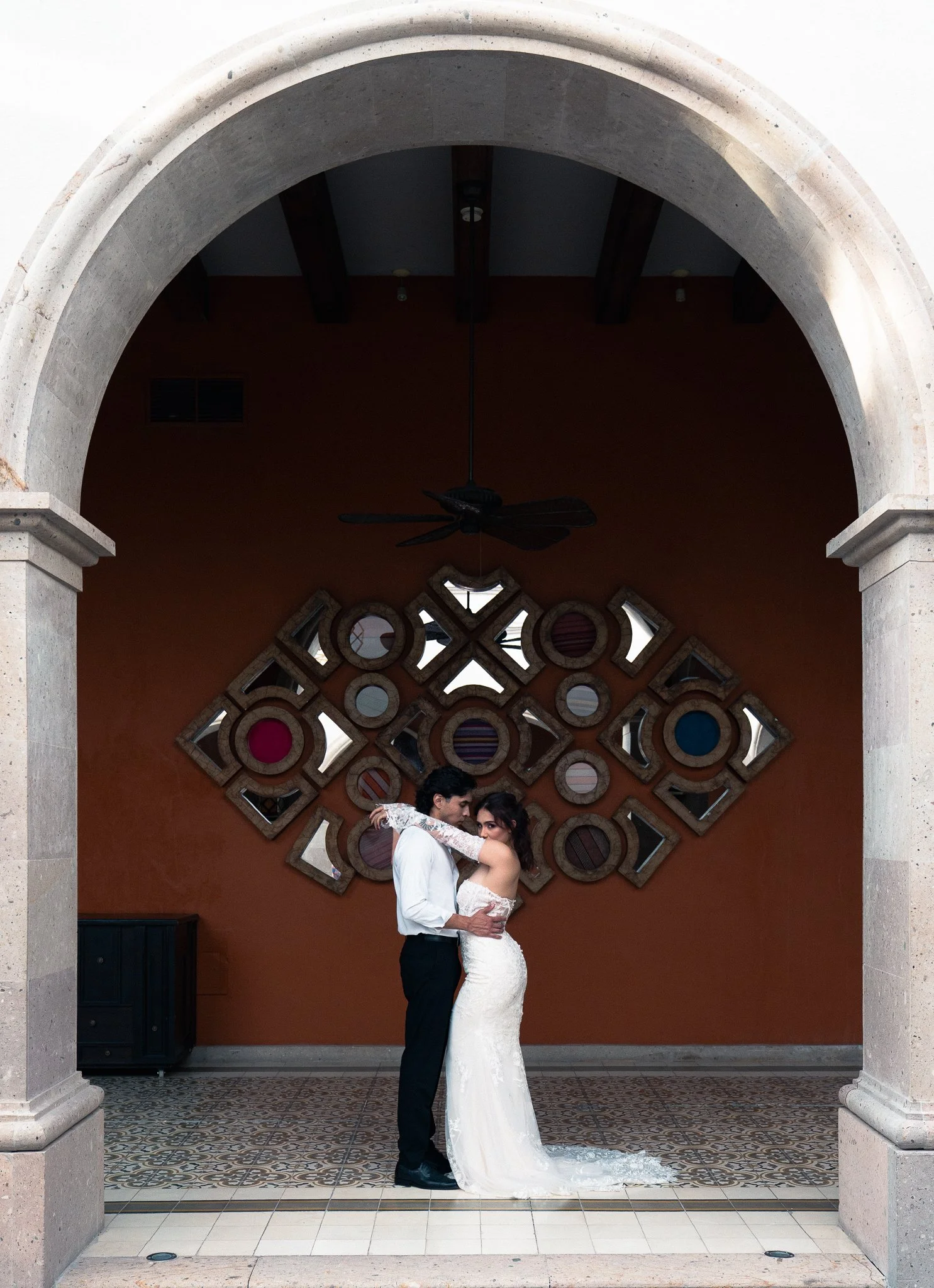 Pareja vestida de boda abrazándose en un arco de piedra, fondo decorativo con espejos y paredes naranja.