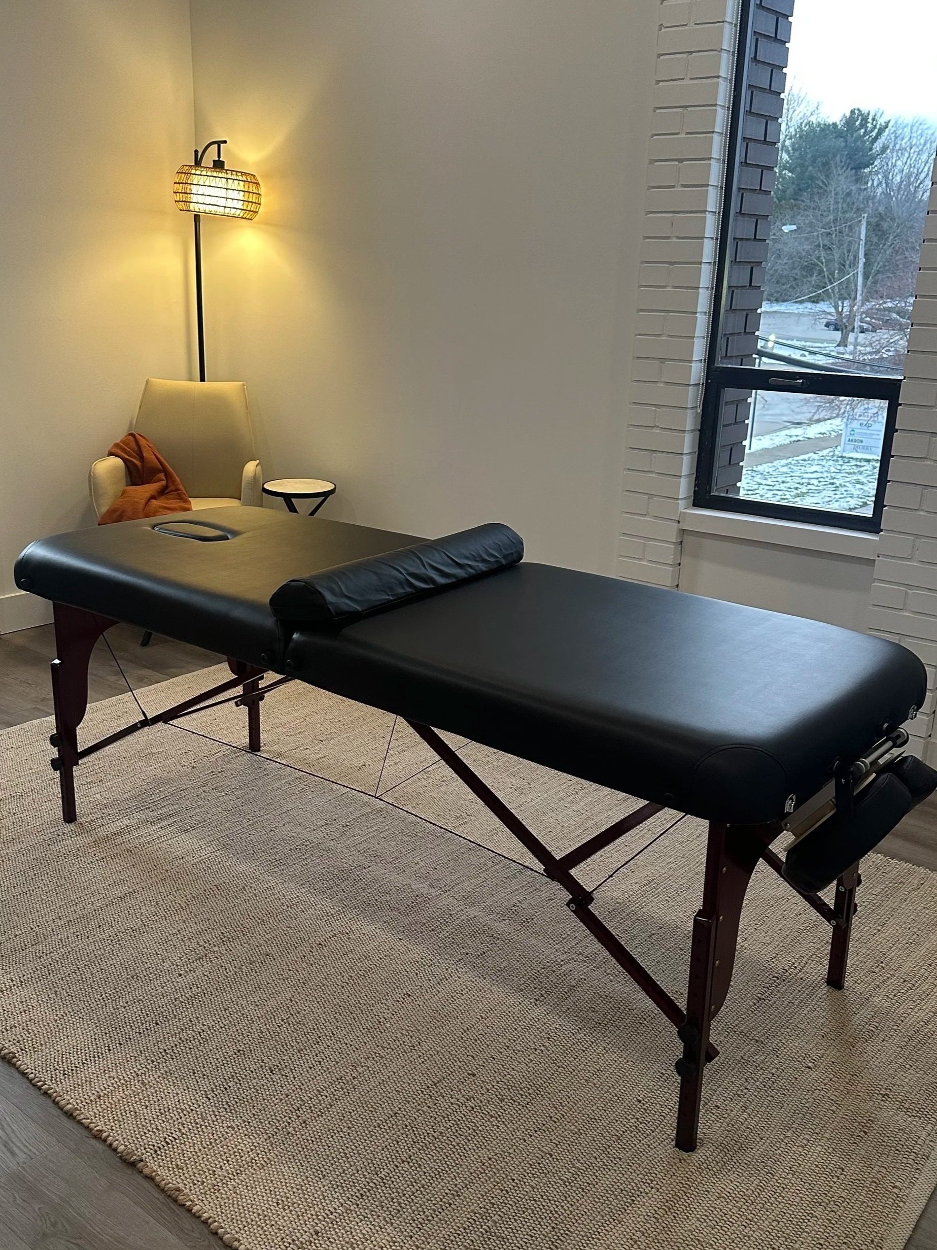 Massage table in a room with a window showing a snowy outdoor scene, a white armchair with an orange blanket, and a standing lamp with a woven lampshade.