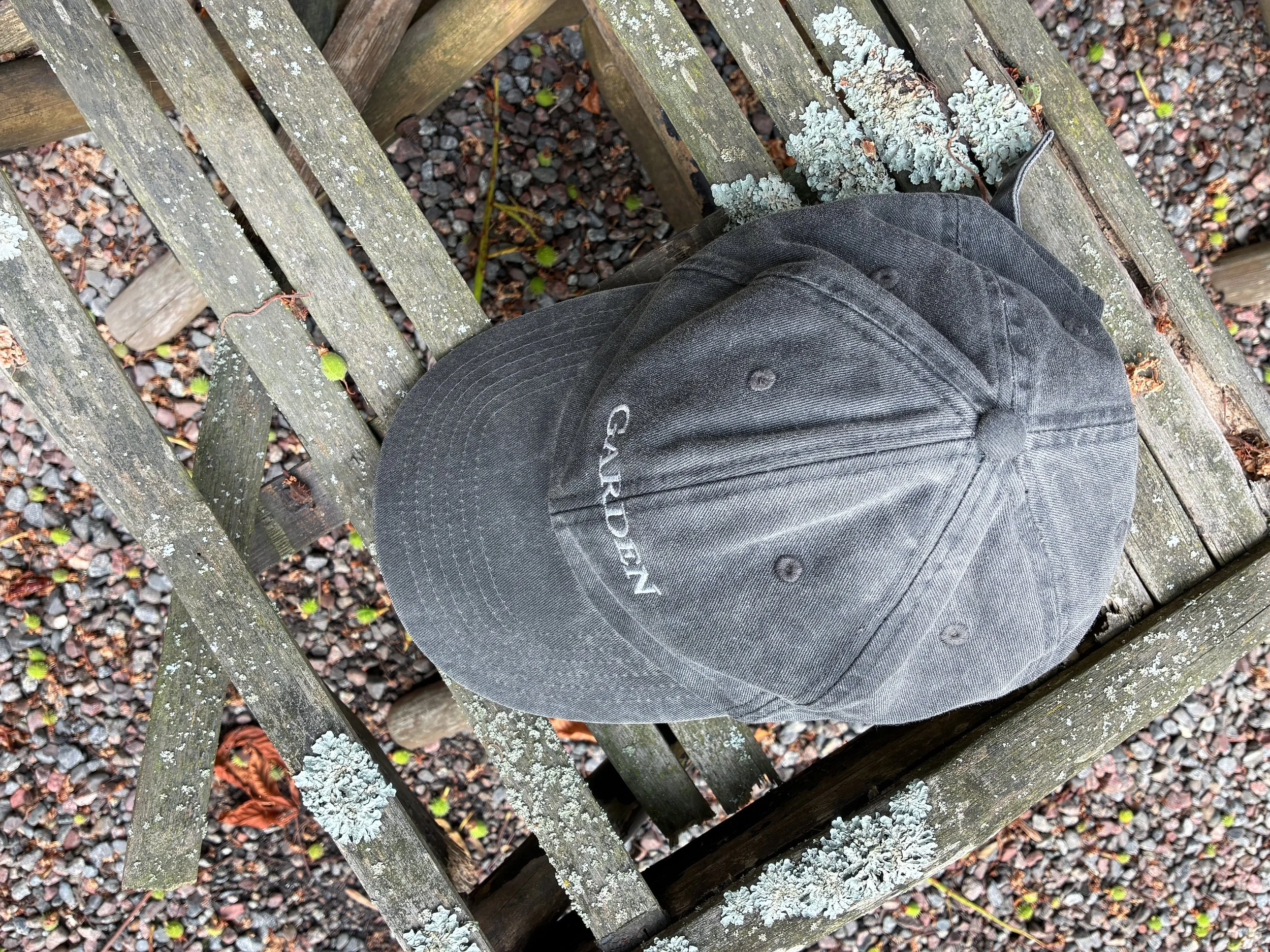 A gray dad cap with the word 'GARDEN' and Tiny Studio embroidered in white, placed on a chair in a garden setting.