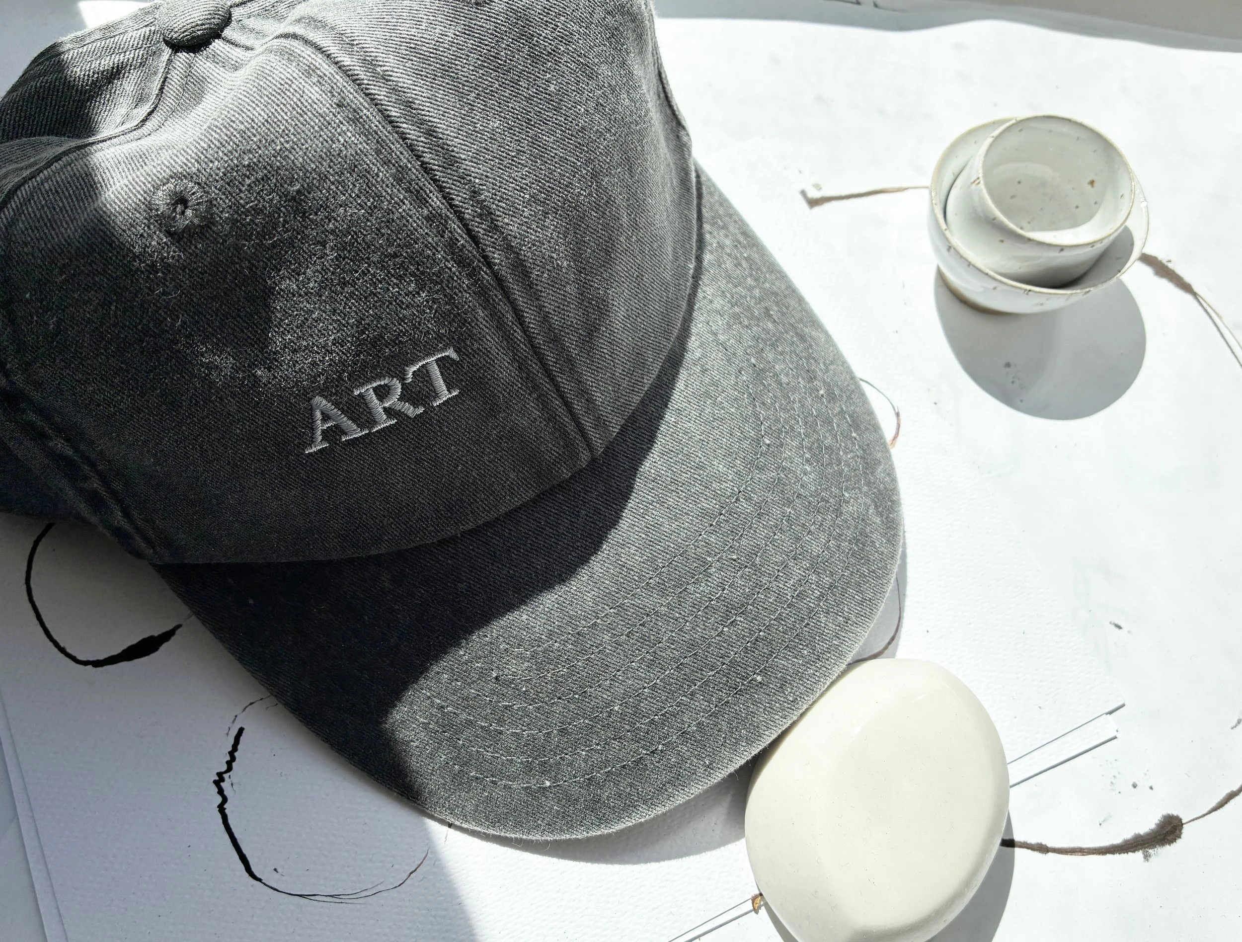 A gray dad cap with the word 'ART' and Tiny Studio embroidered in white, placed on a white surface with black lines, next to three white mugs stacked together.