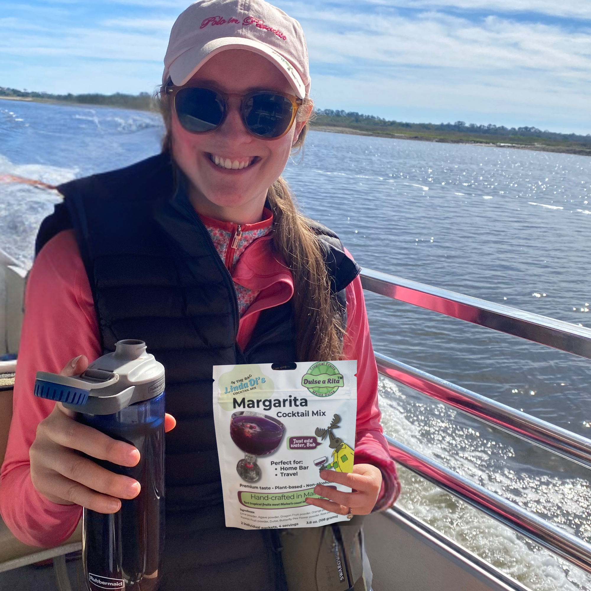 A woman smiling on a boat holding a Margarita cocktail mix packet and a water bottle, with water and a distant shoreline in the background.