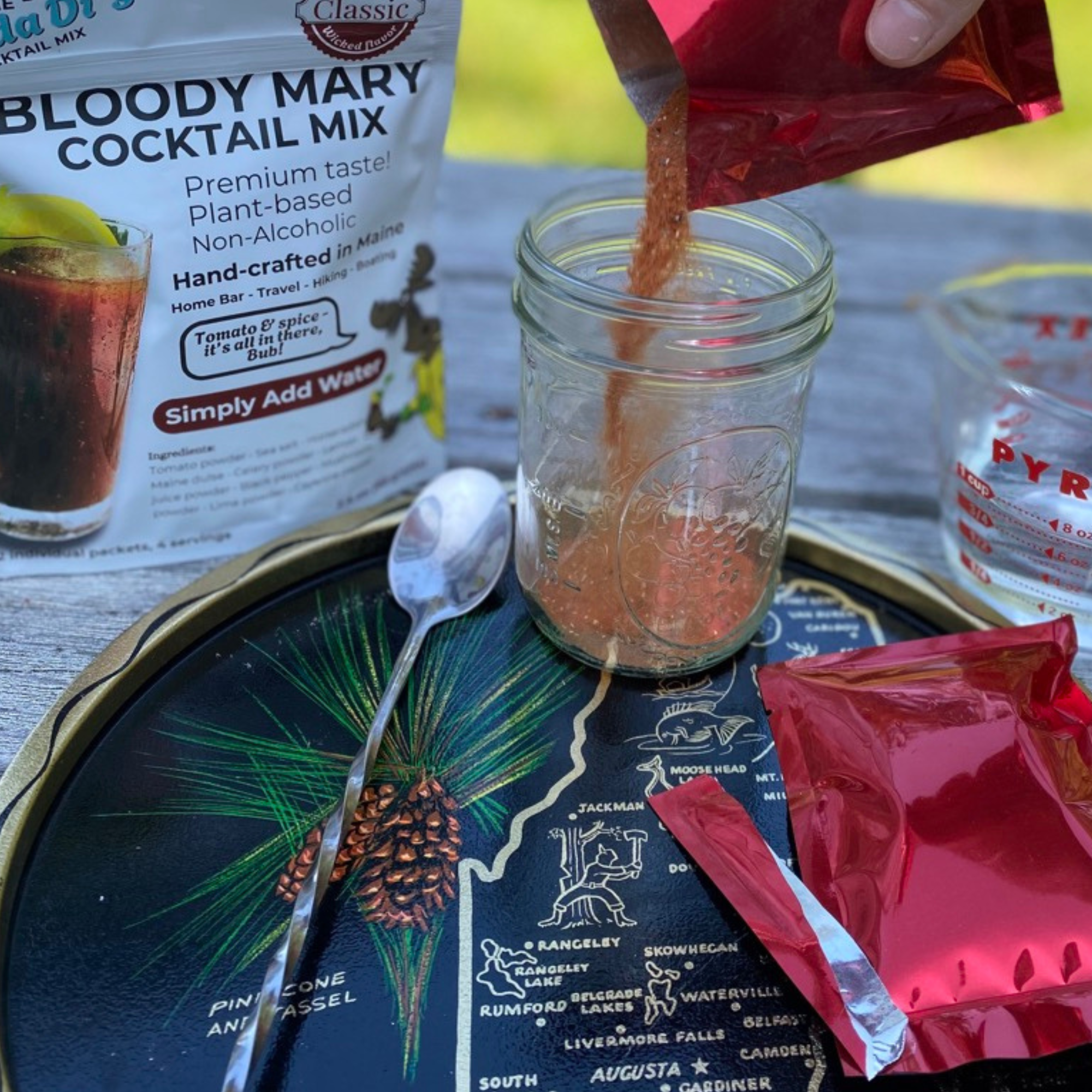 A bag of Bloody Mary cocktail mix being poured into a mason jar with a Spoon, surrounded by a measuring cup and red packets, placed on a decorative tray with a map of Maine.