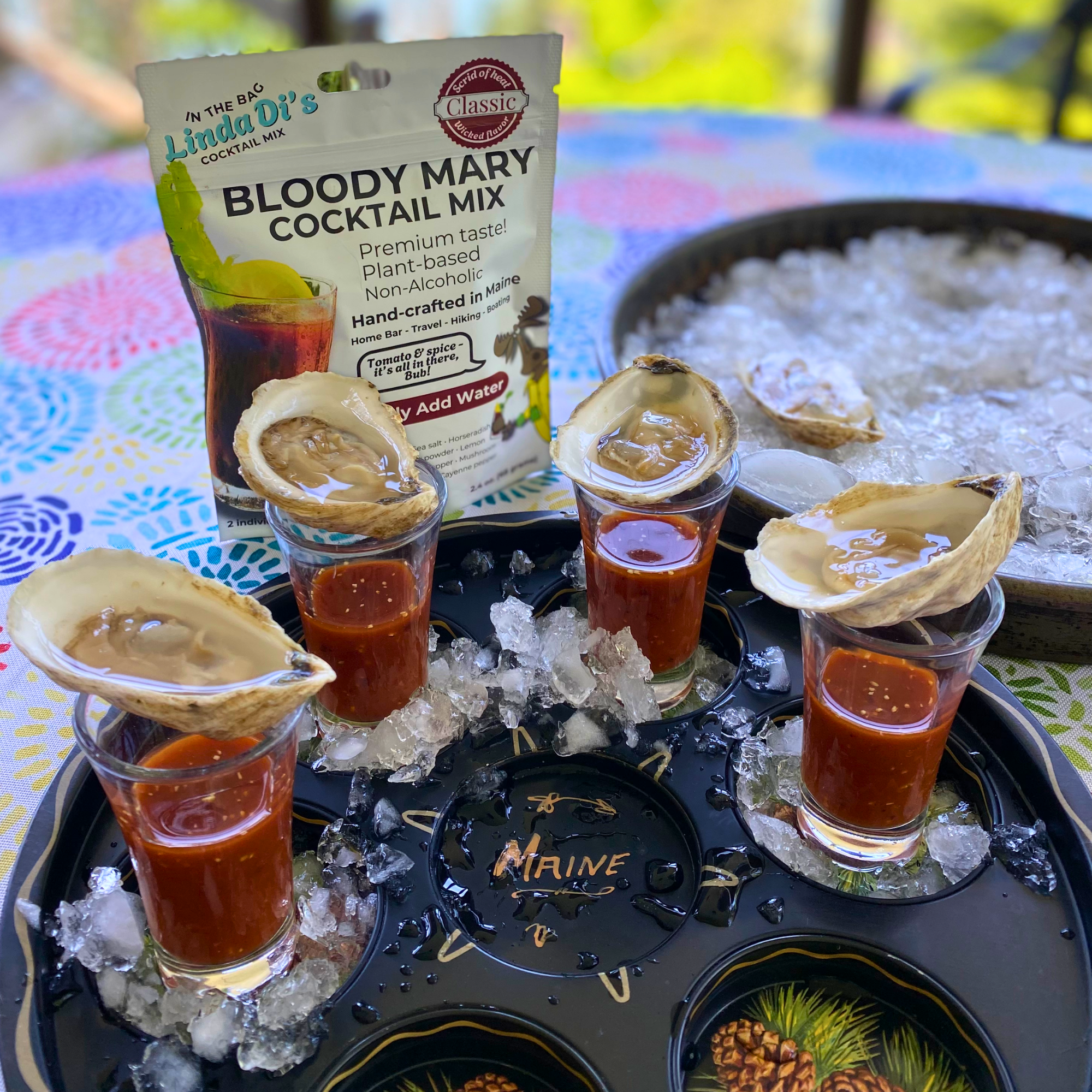 Six shot glasses with Bloody Mary cocktail garnished with oyster shells, ice on a tray with the word 'MAINE' on it. In the background is a bowl of ice and a packet of Lindad's Bloody Mary cocktail mix.