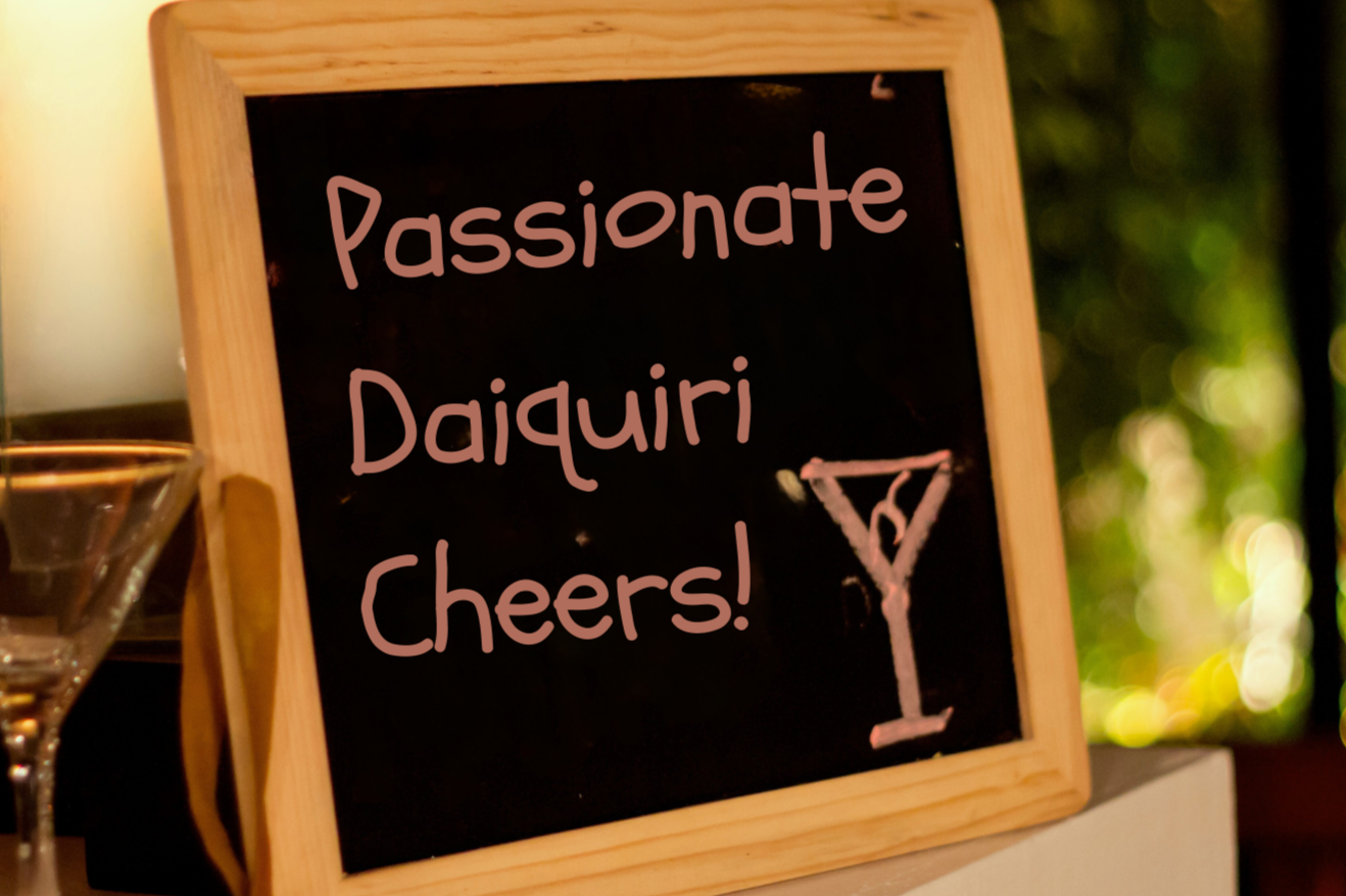 A framed blackboard sign with the message 'Passionate Daiquiri Cheers!' and a drawing of a martini glass. The sign is placed on a table near a glass with a lemon wedge, with a blurred outdoor background.
