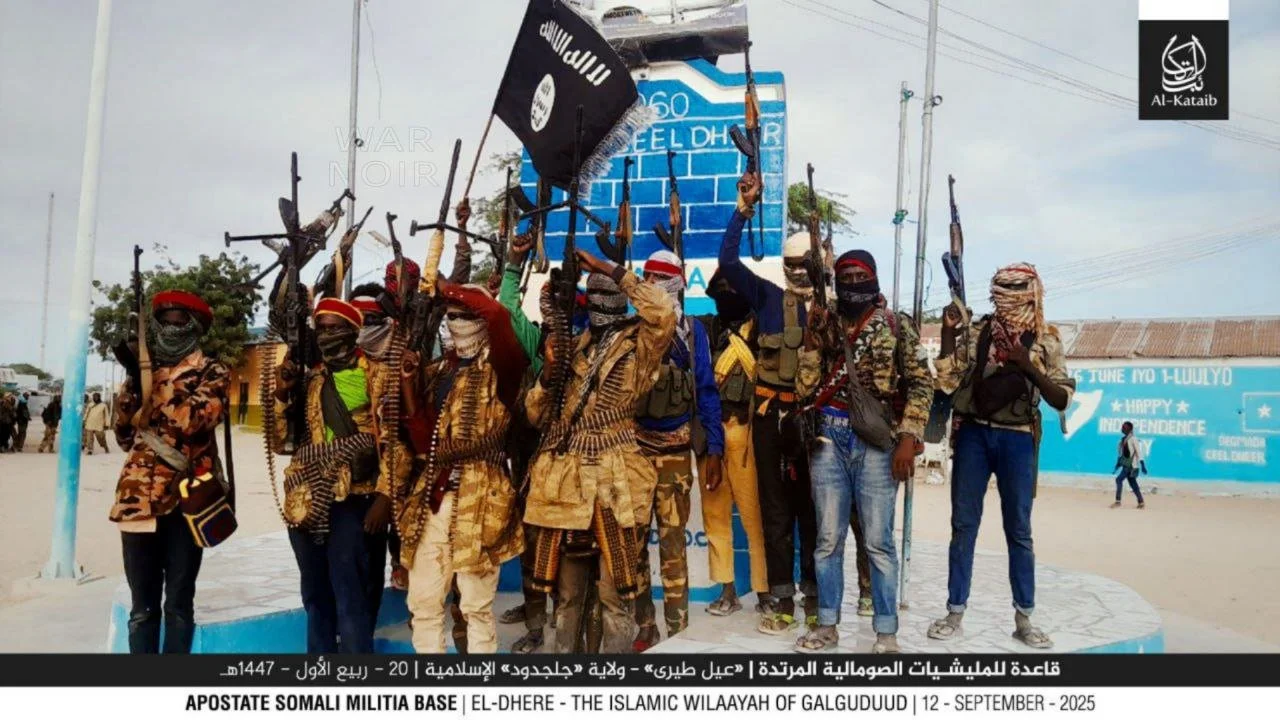 Group of armed militants holding rifles and a black flag with Arabic writing in an outdoor setting