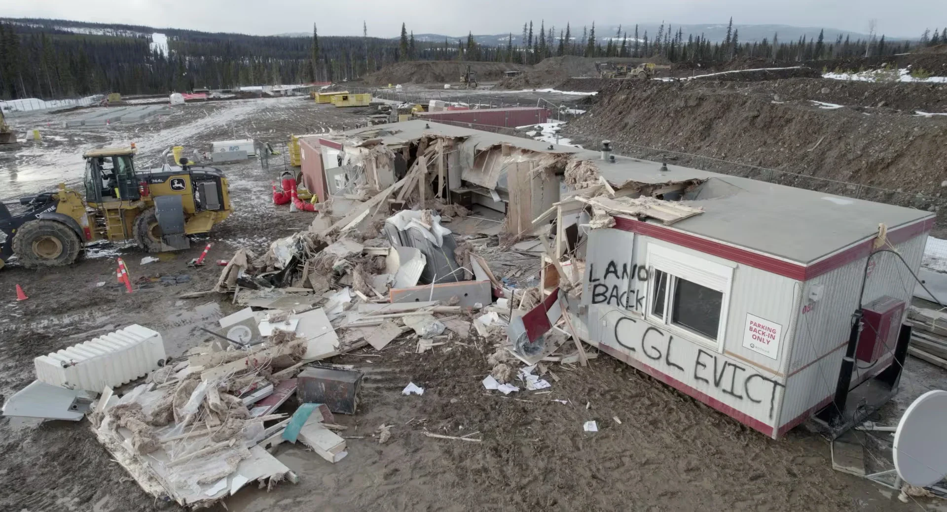 Damaged trailer structure and debris at a cleared site with construction equipment in a rural area