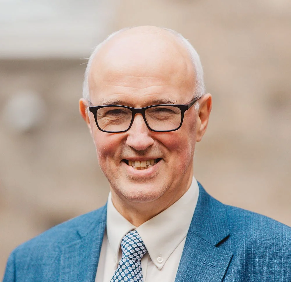 Bald man wearing glasses and a blue suit with a patterned tie, smiling.