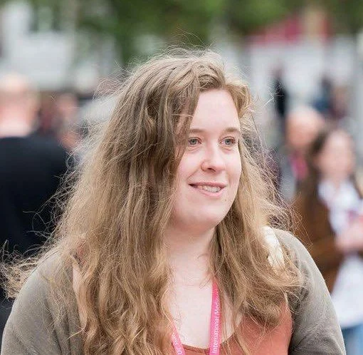 Woman with long hair wearing a pink lanyard, outdoors, with people in the blurred background.