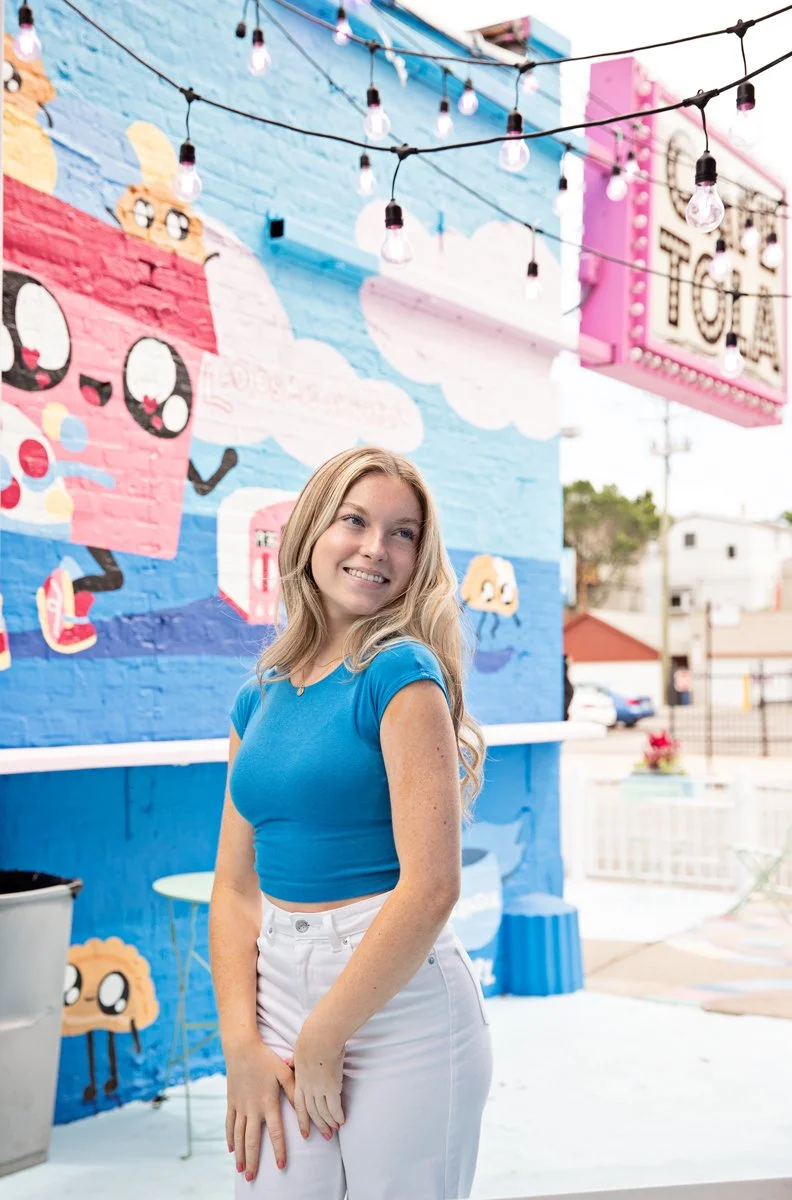 A young woman with blonde hair wearing a blue crop top and white pants standing in front of a colorful mural of a cartoon ice cream cone. The mural includes clouds and other playful designs. String lights are hanging overhead.