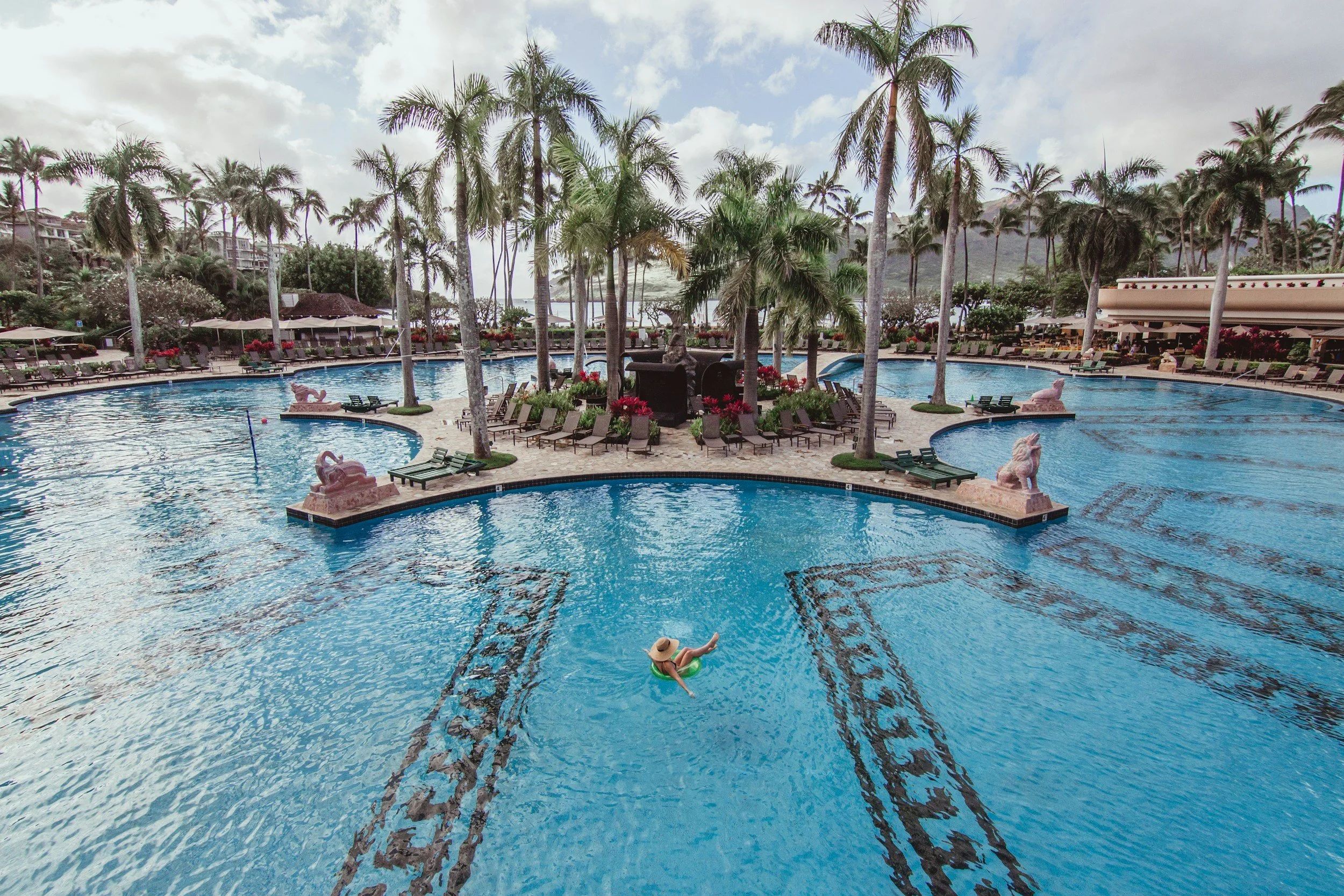 Pool area with palm trees, lounge chairs, umbrellas, and sculptures, with a person floating on a pool float and mountains in the background.