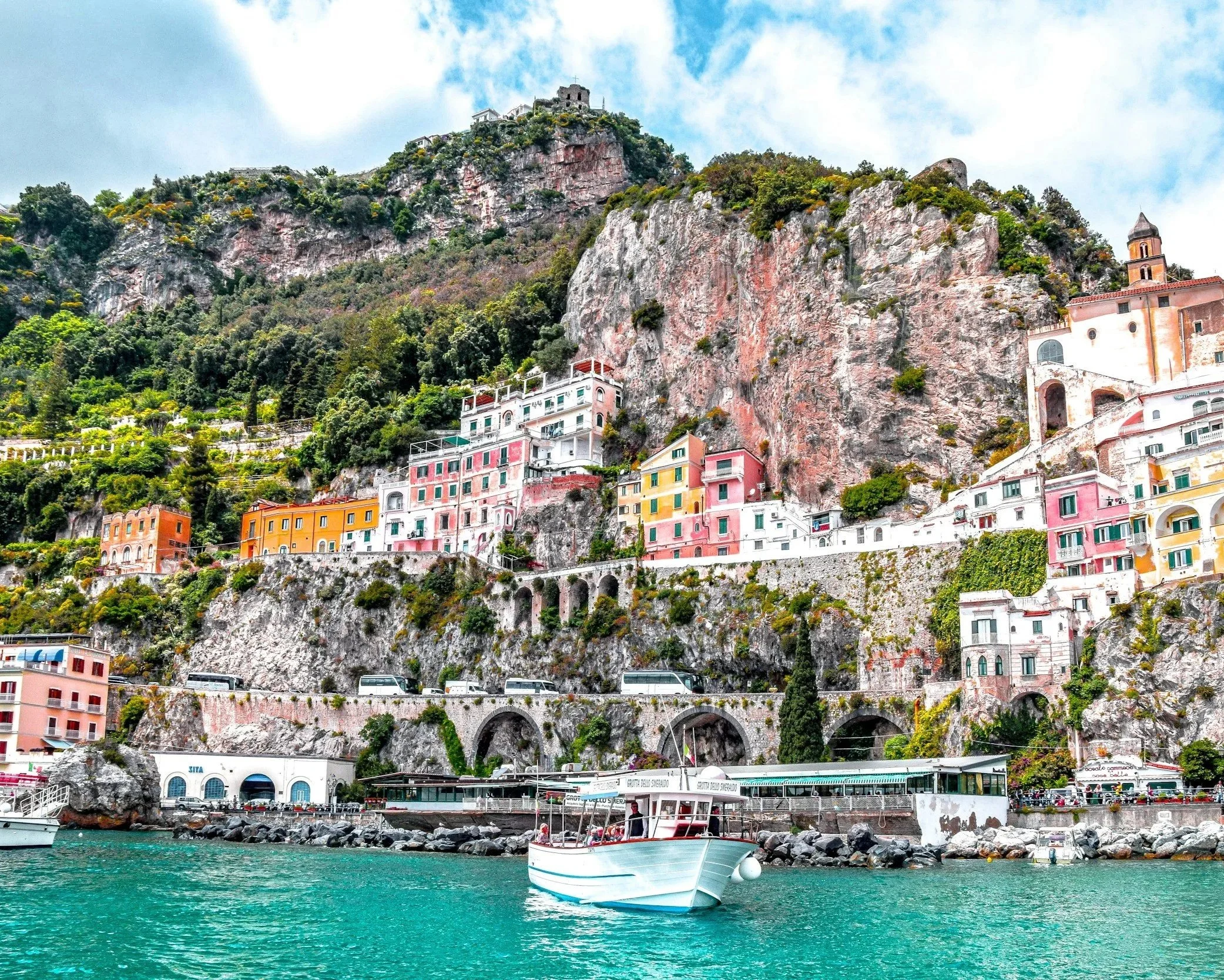 Colorful buildings and a church built into a hillside with lush greenery, overlooking a harbor with boats, in a coastal town with a castle on top of the hill.