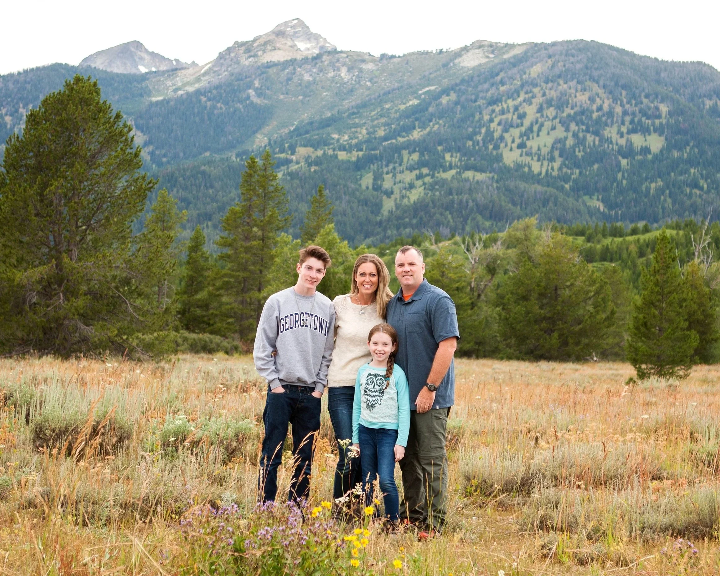 A family of five standing outdoors in a meadow with wildflowers, trees, mountains, and a cloudy sky in the background.