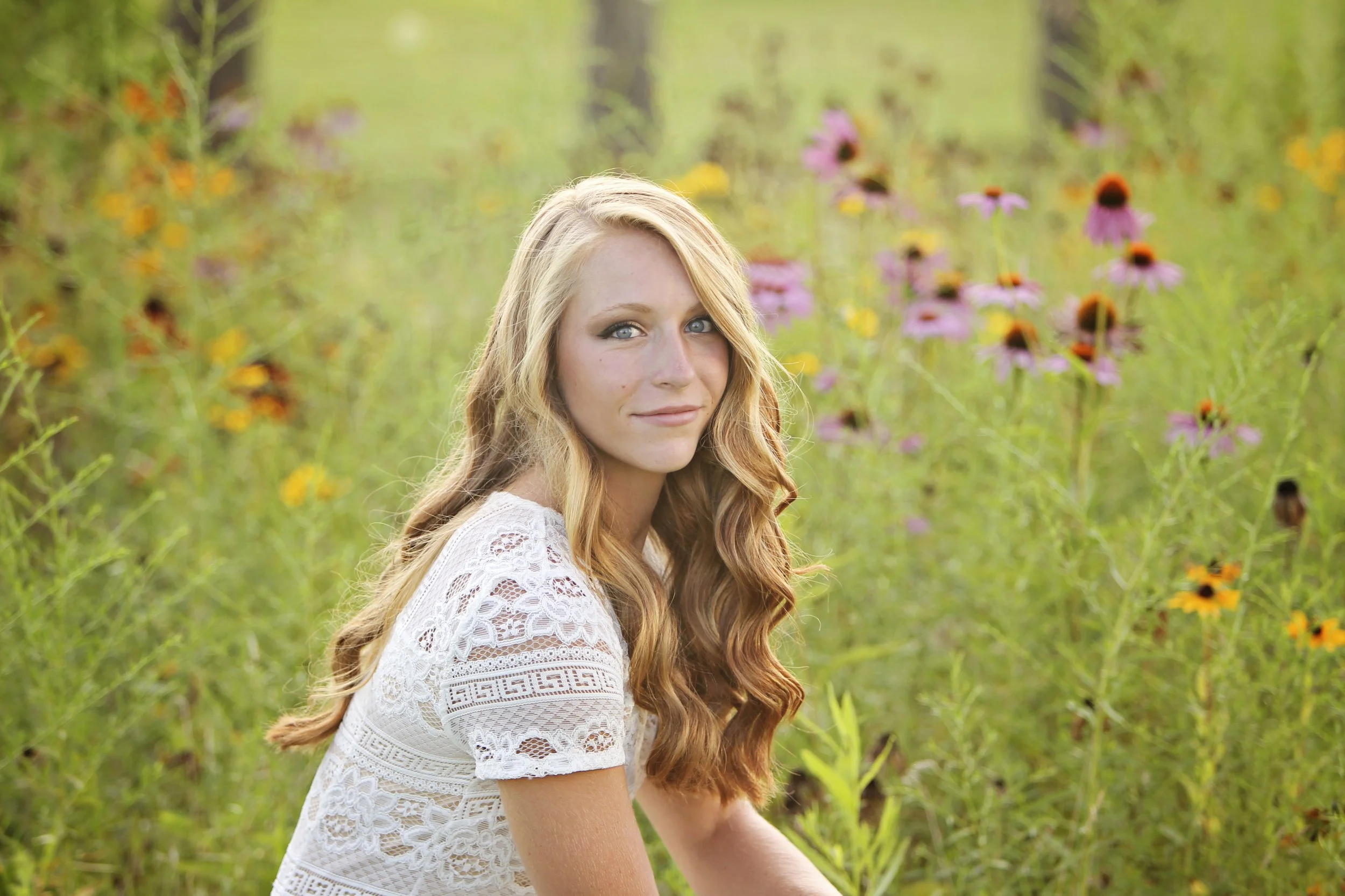 A young woman with long red hair and blue eyes sitting in a field of pink, orange, and yellow wildflowers on a sunny day.