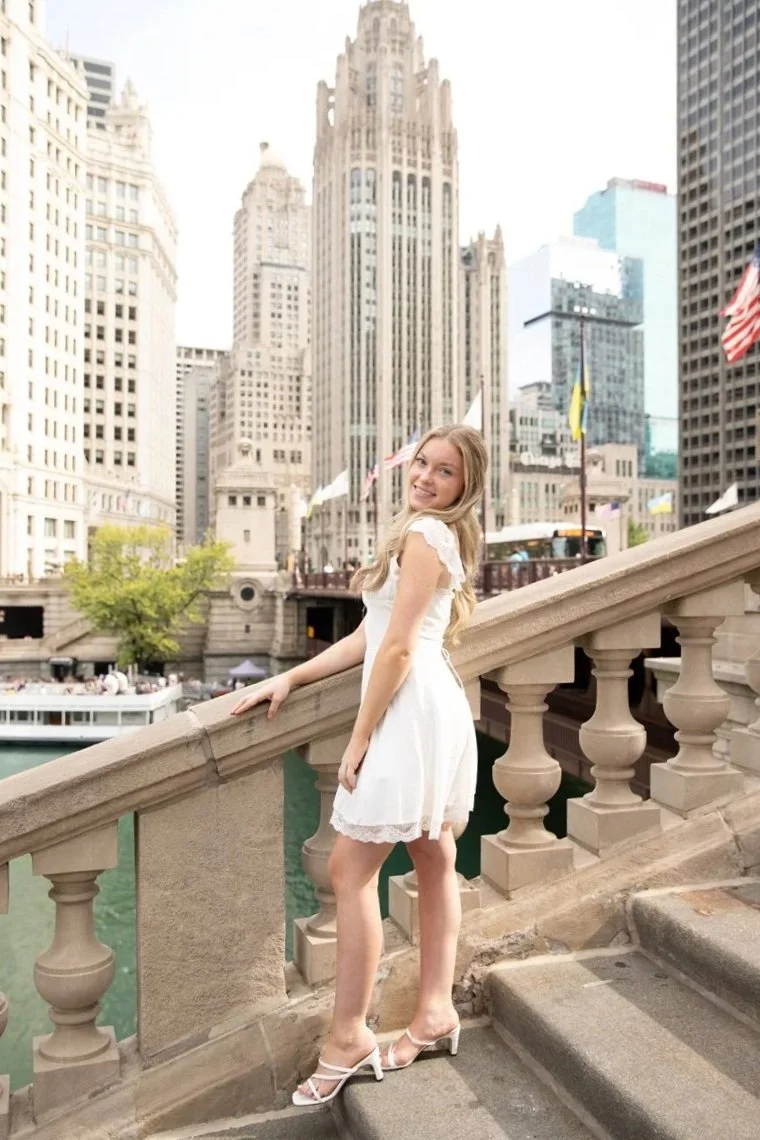 A young woman in a white dress and high heels smiling and leaning on a bridge railing with a cityscape of tall buildings in the background, including the Wrigley Building and Chicago River.