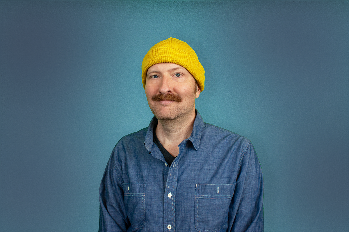 Young man with a mustache wearing a yellow knit beanie, denim shirt, and a black T-shirt underneath, standing against a blue background.
