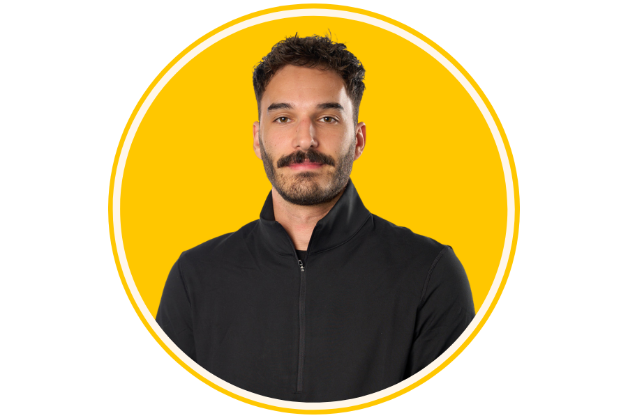 A man with dark hair, a mustache, and light beard, wearing a black t-shirt with white text, standing against a yellow background with a circular white outline.