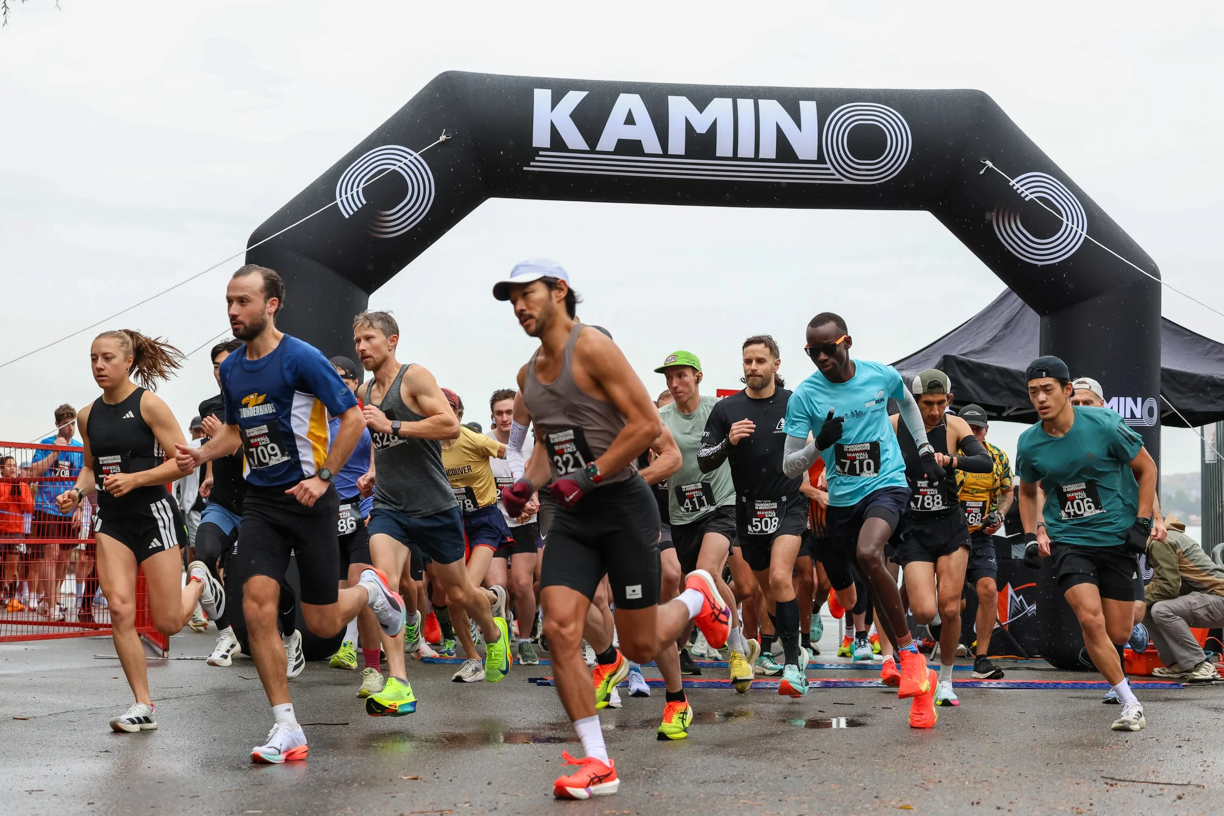 Marathon runners starting race under a large black arch with the word 'KAMINO' above it, on a wet street, with some spectators and tents in the background.