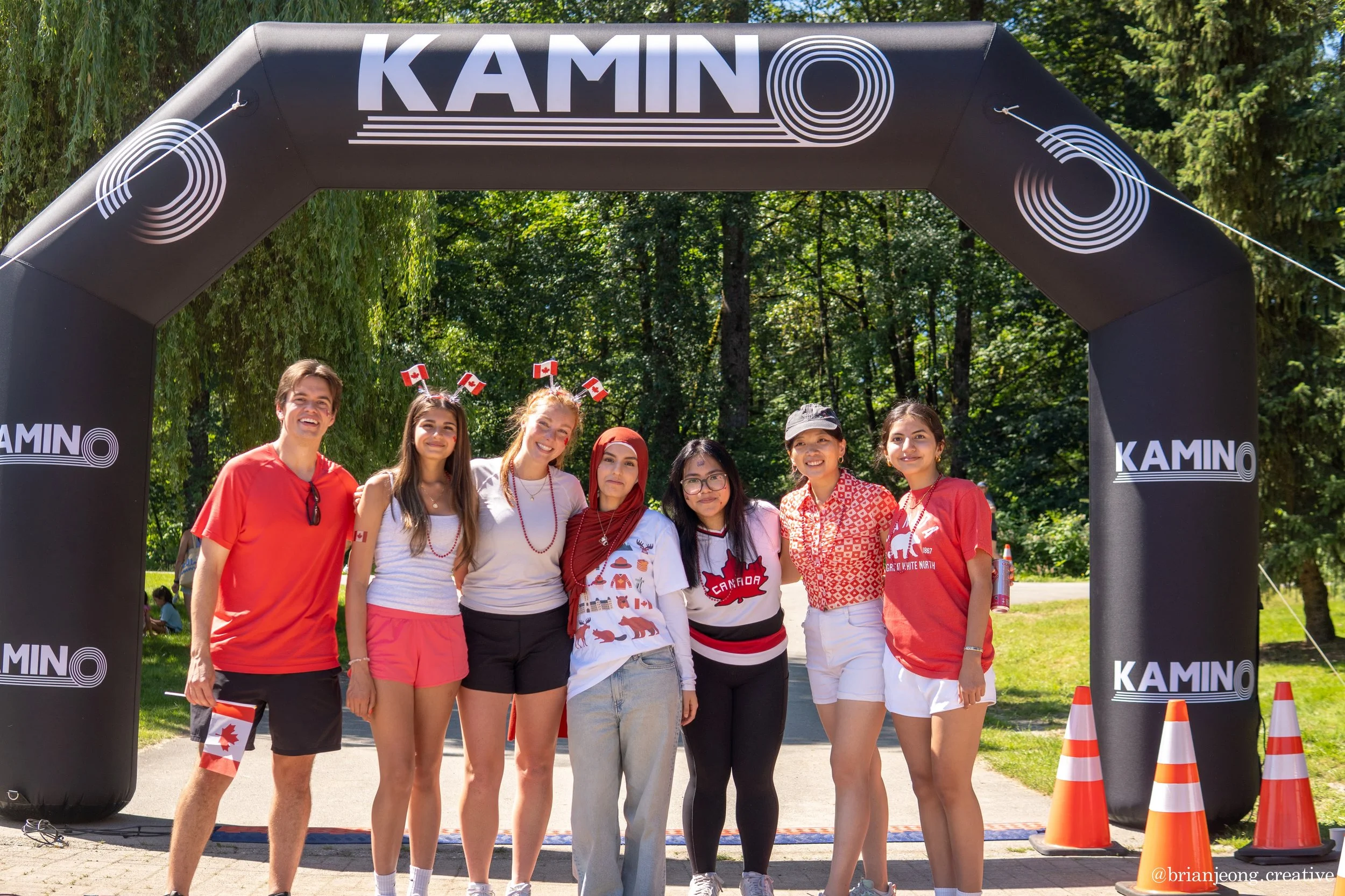 Group of seven young people standing under an inflatable arch with the word 'KAMINO' in a park, dressed in red and white clothing, some with Canadian flags, smiling at the camera.