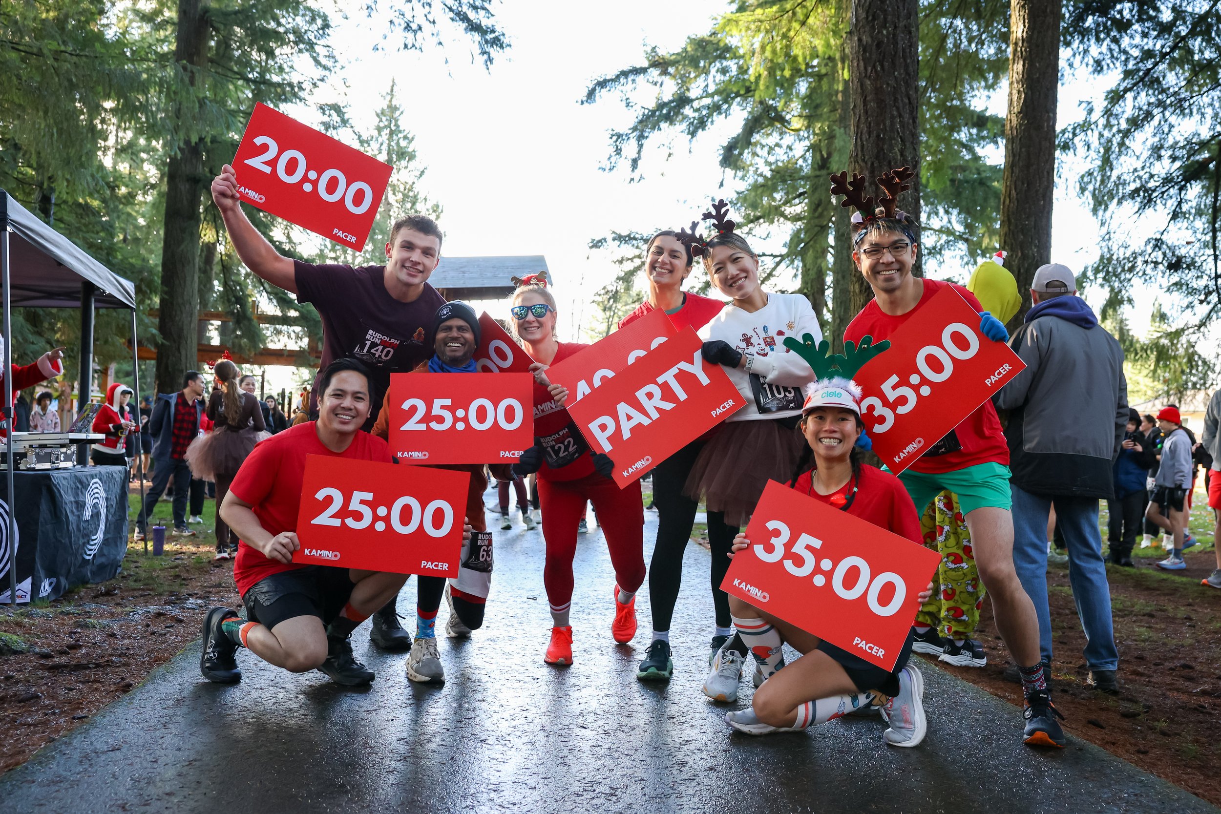 Group of runners celebrating after a race, wearing holiday-themed accessories, holding signs with finish times and a party sign, in a wooded outdoor park setting.