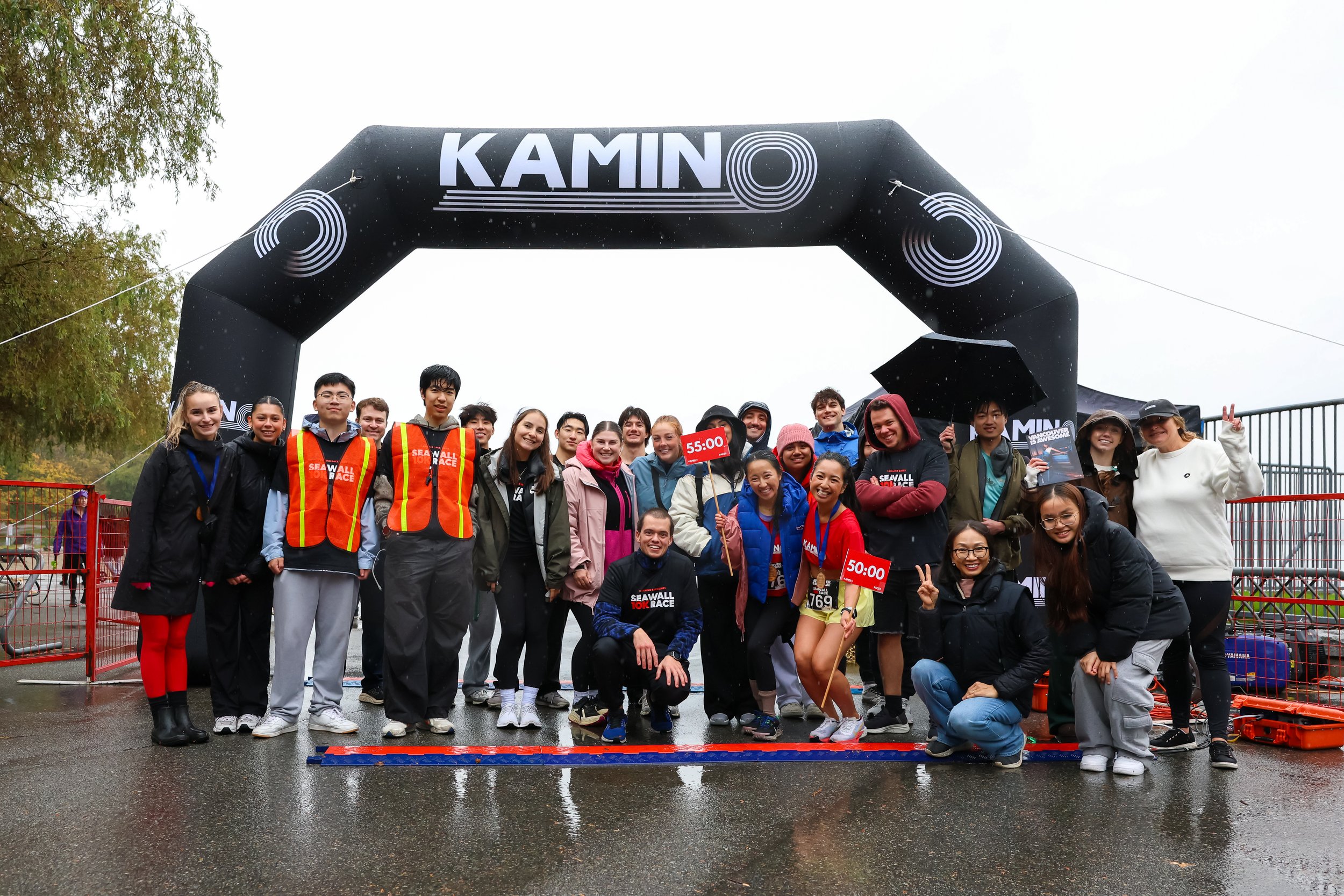 Group of people celebrating at a rainy outdoor race event under a large black arch with the word 'KAMINO' on it. Some are holding signs with race times, and others are making peace signs or smiling.