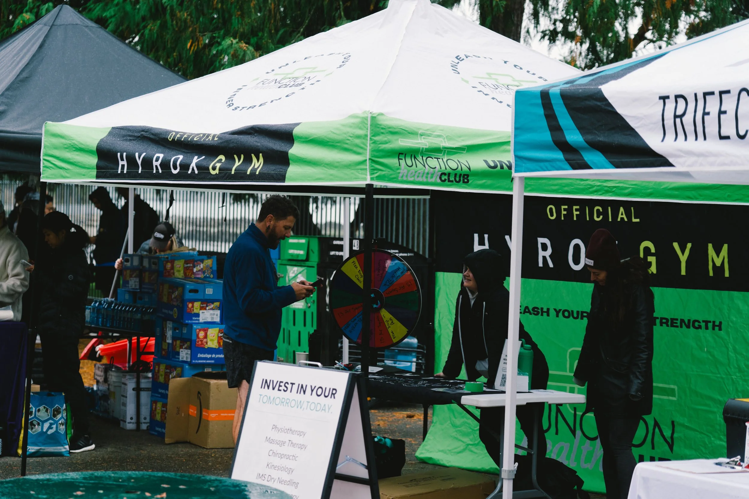 Outdoor event with a tent labeled 'Official Hyrokgym' and people around it, including a man looking at his phone, a woman smiling, and a woman in a beanie, with a spinning wheel and display table.