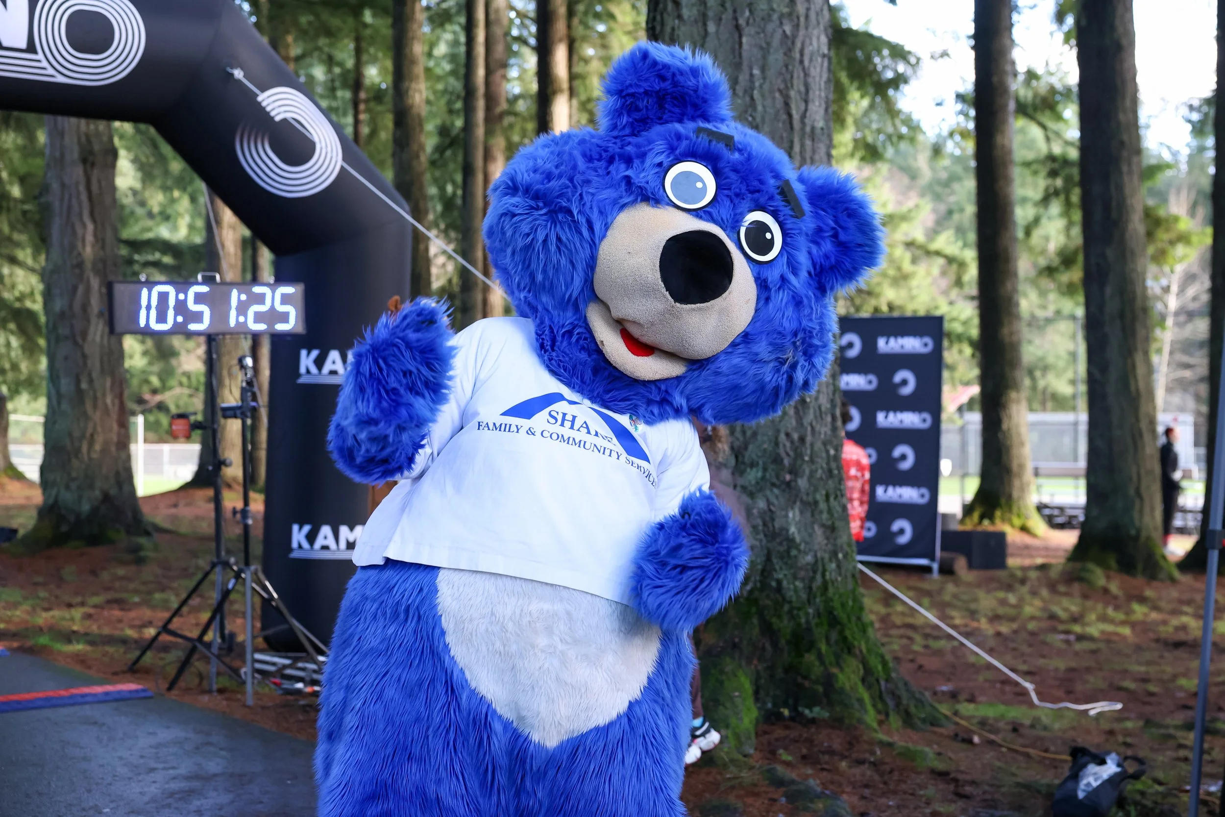 Blue bear mascot wearing a white T-shirt with blue text, standing outdoors among trees, with a digital clock and event banners in the background.