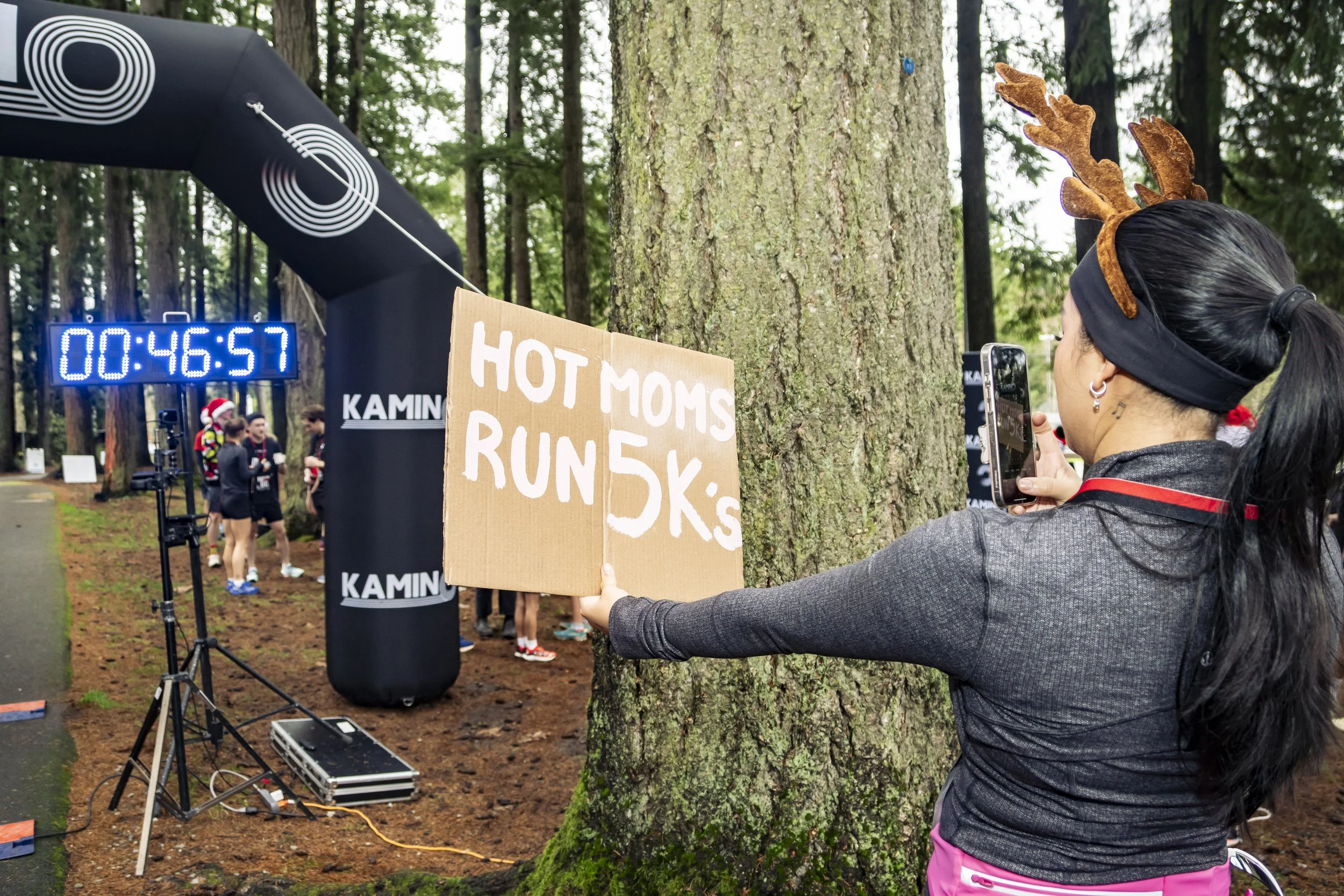 Woman with reindeer antler headband taking a photo with her phone at a running event in a wooded area, holding a cardboard sign that says "HOT MOMS RUN 5K's." There is a digital timer showing 46 minutes and 57 seconds and runners near the start/finis