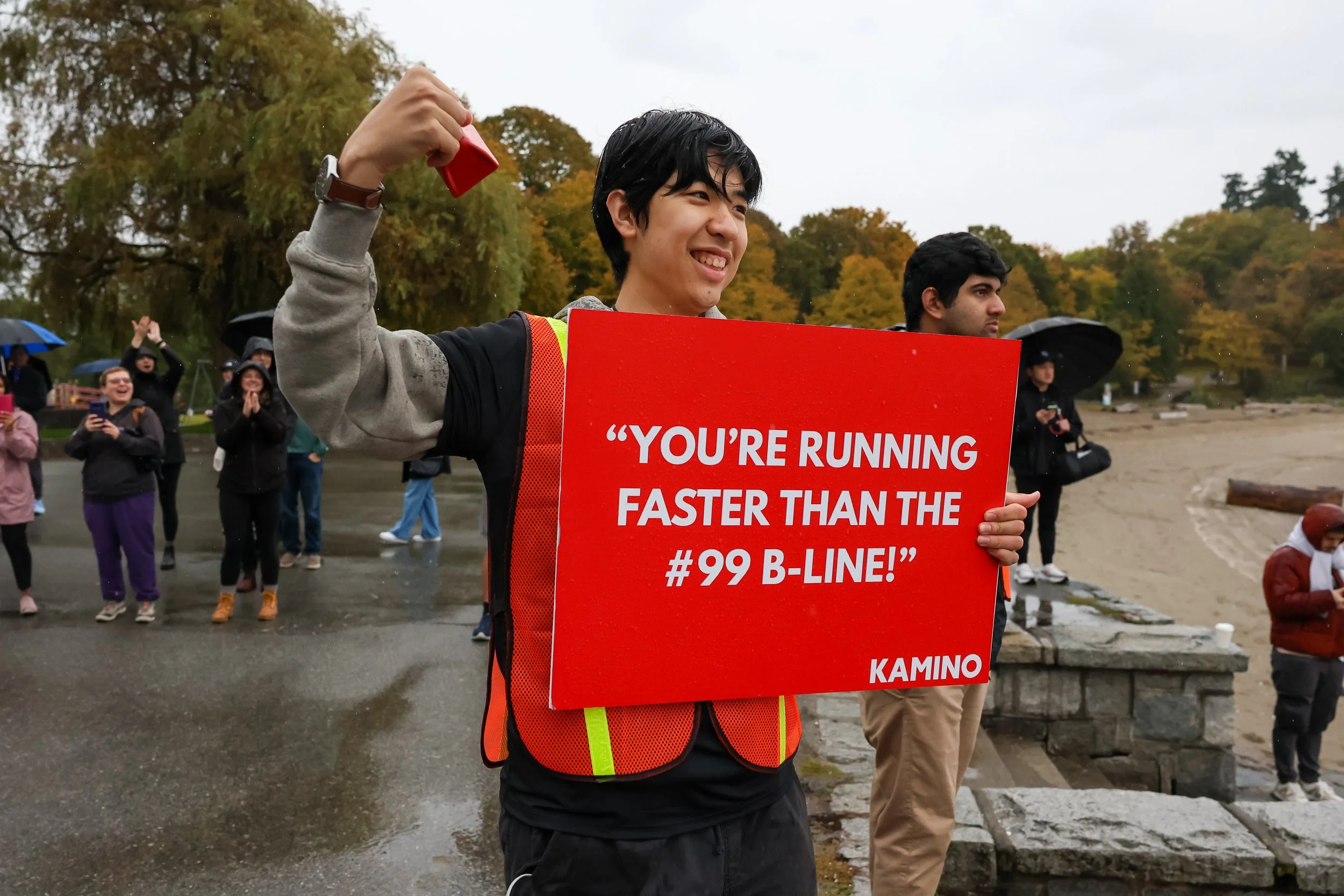 A young man holding a red sign that reads, "You're running faster than the #99 B-Line!" at an outdoor event with other people in the background, some holding umbrellas, on a cloudy, rainy day near a beach.