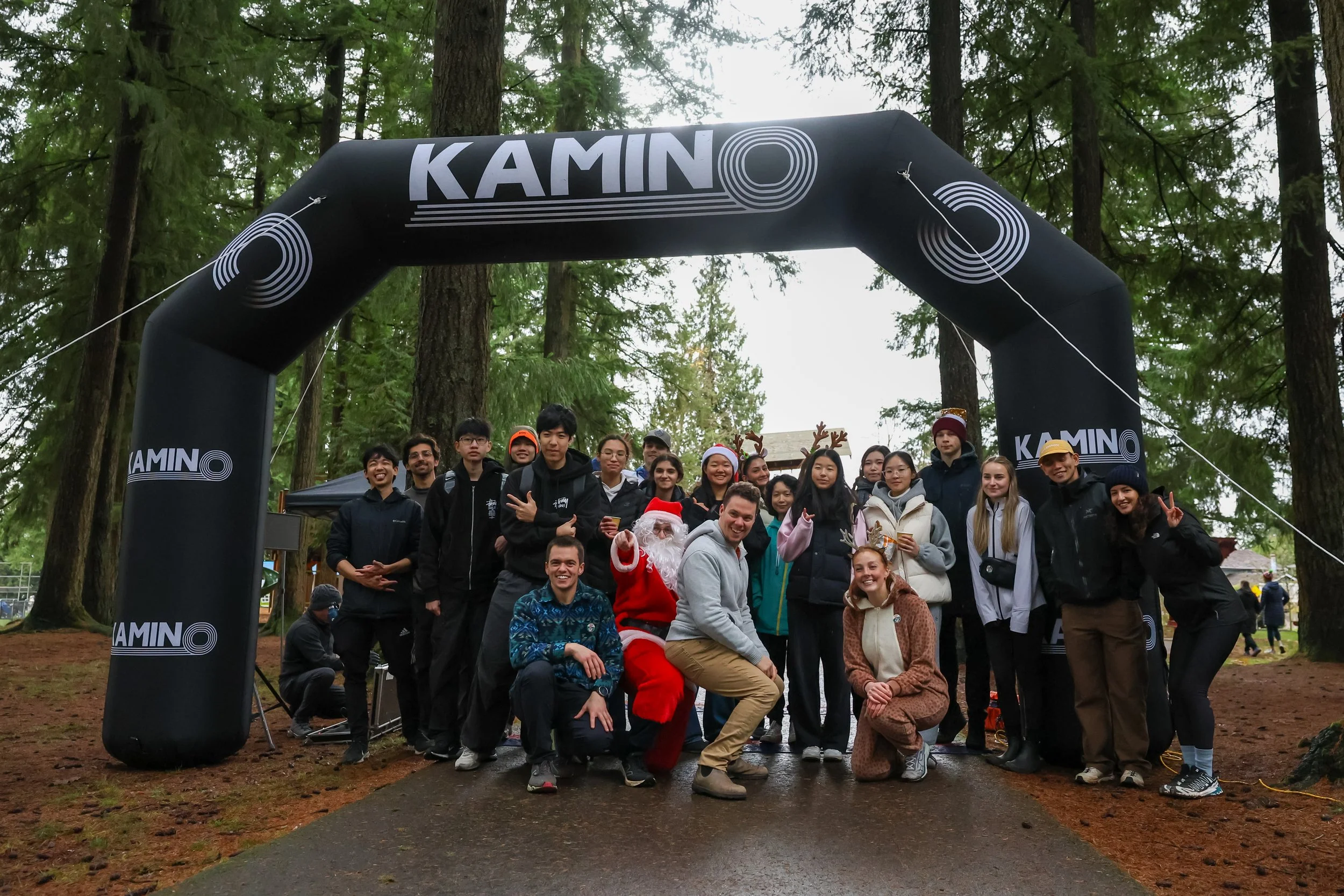 Group of people posing under a black inflatable archway with the word 'KAMINO' printed on it, in a wooded outdoor area with tall trees, some wearing festive costumes and reindeer antlers.