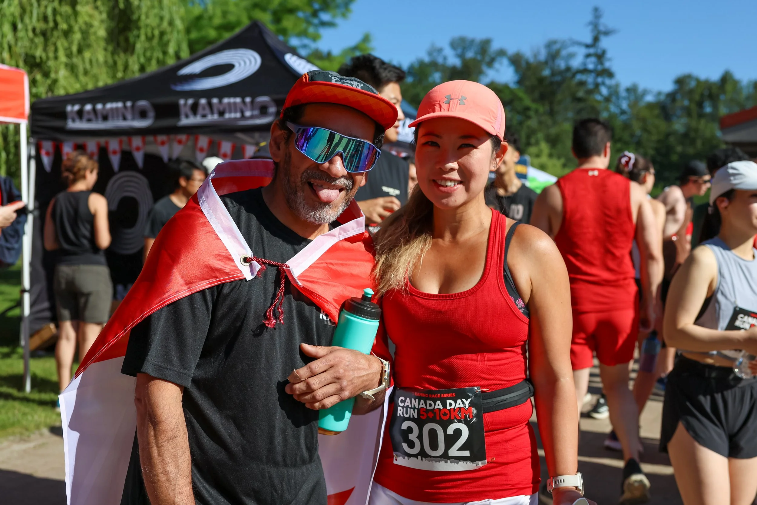 Two runners at a race event, one man and one woman, smiling and posing for the photo, with a crowd and race tents in the background.