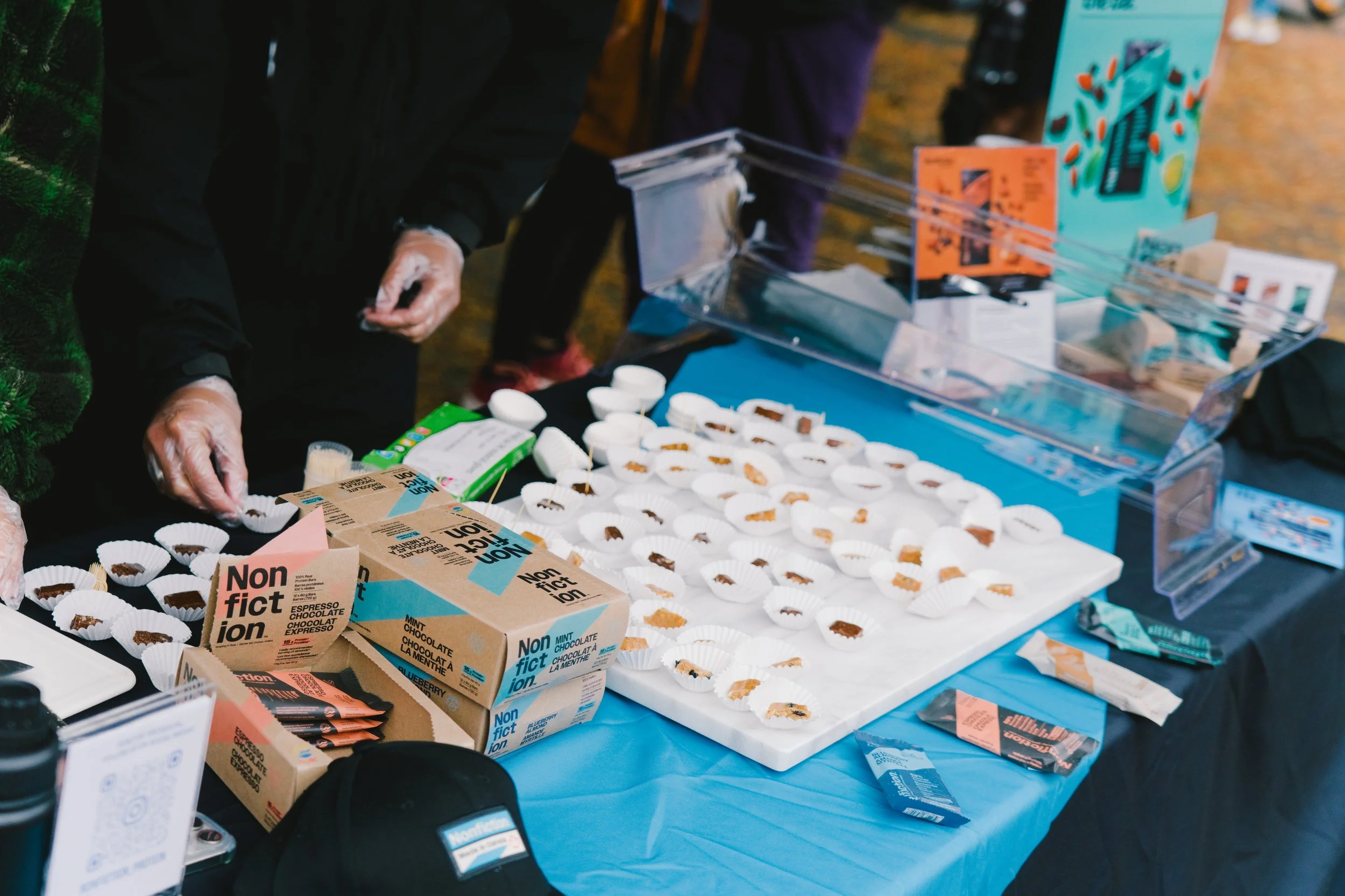 Display table at a public event with chocolates and informational cards, with people's hands wearing gloves preparing samples.