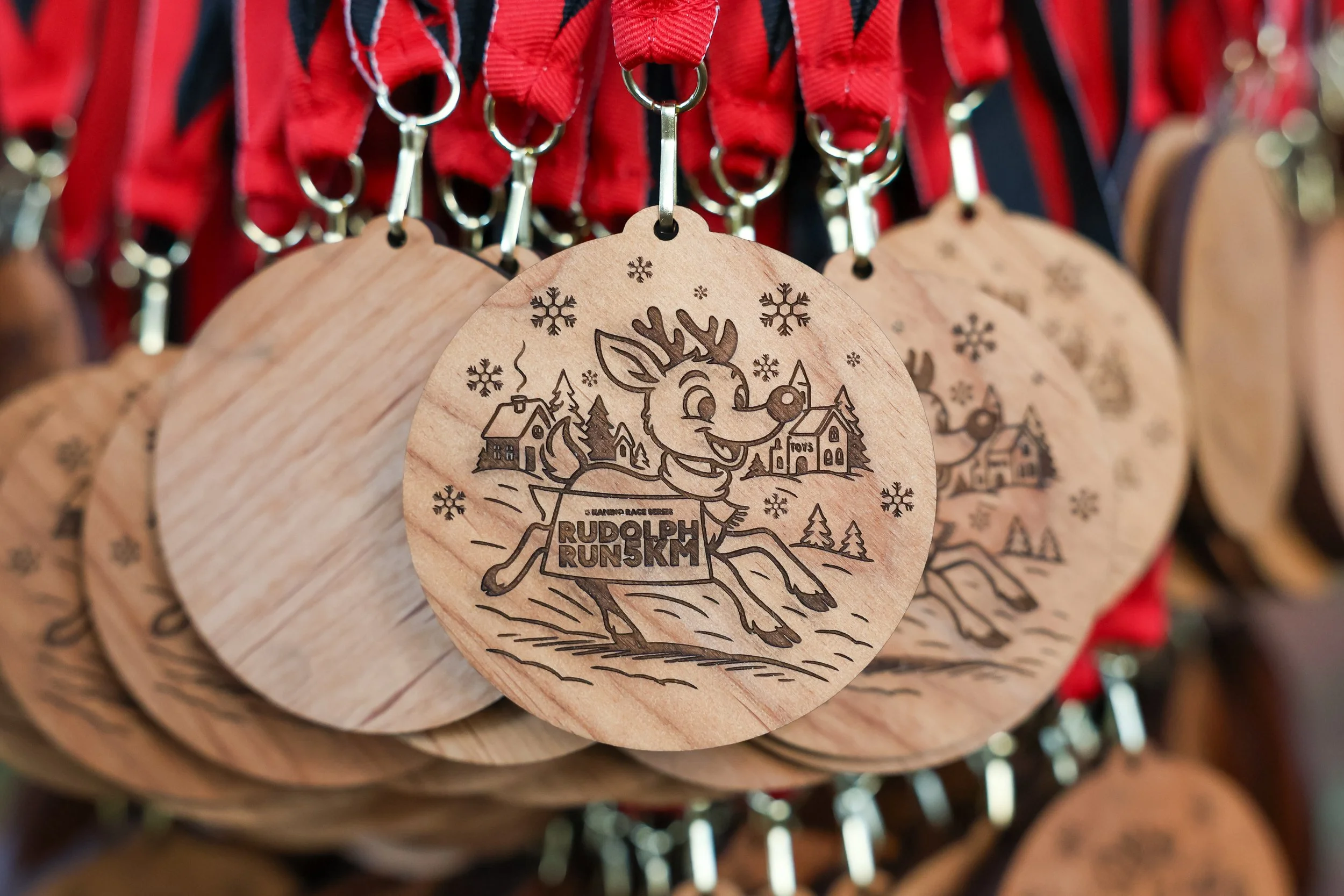 Wooden keychain with an engraved illustration of Rudolph the Red-Nosed Reindeer running, surrounded by snowflakes and winter scenery. Text on the keychain reads 'Rudolph Run 5K'.
