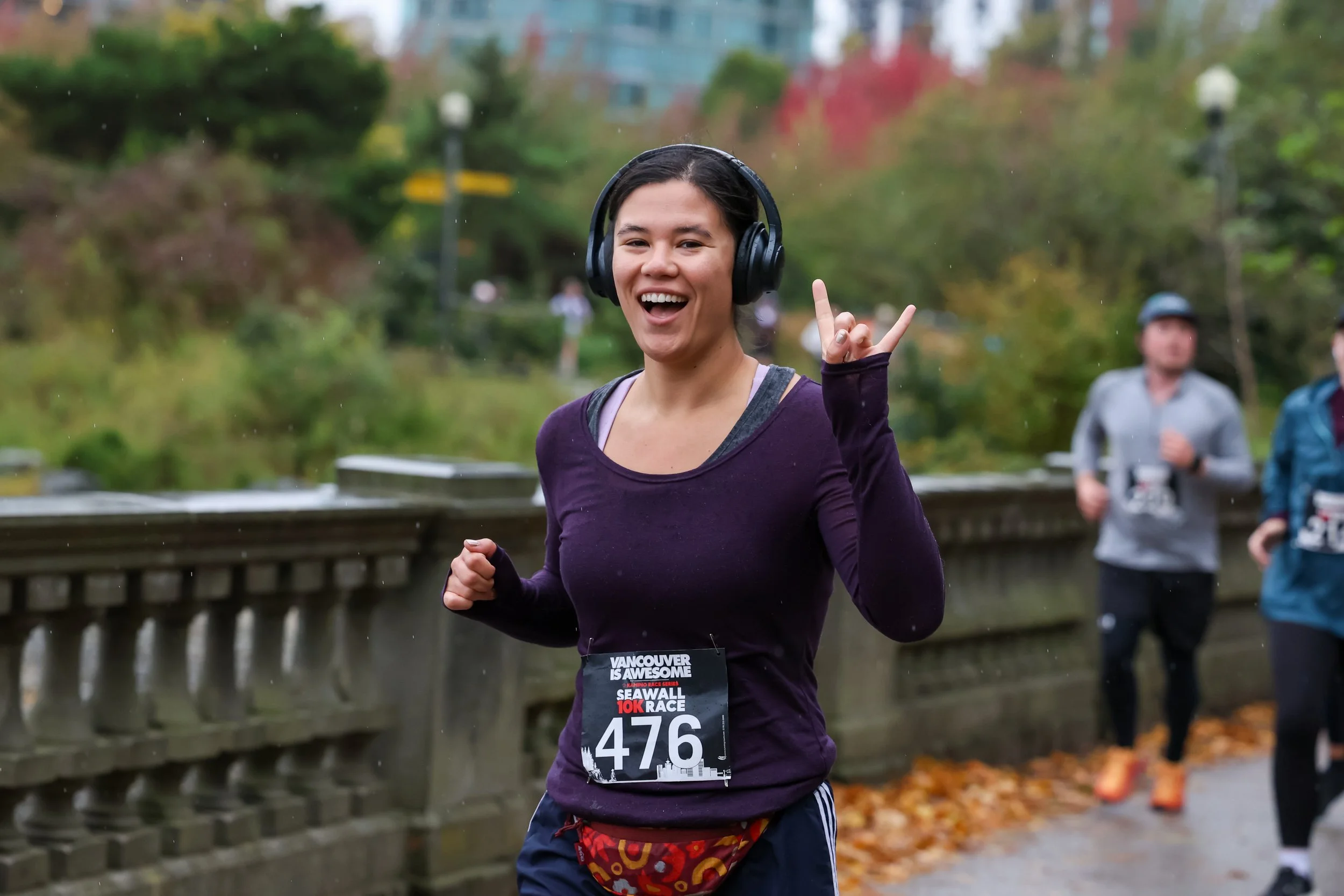 Young woman smiling and running in a race wearing headphones, with race bib number 476, in an outdoor park setting with other runners and autumn foliage.