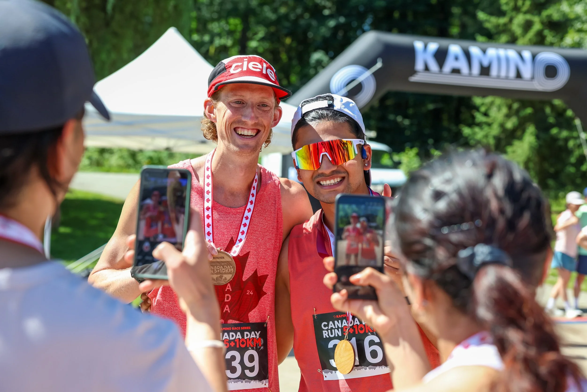 Two runners in red shirts with medals pose for photos at a race event, surrounded by people taking pictures, outdoors with green trees in the background.