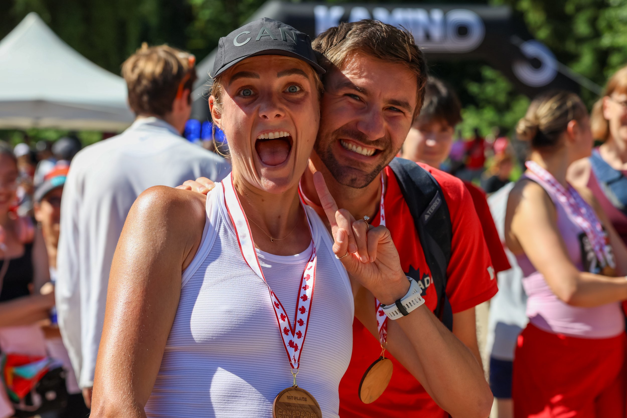 Two happy runners with medals celebrating at an outdoor event, surrounded by crowd and tents.
