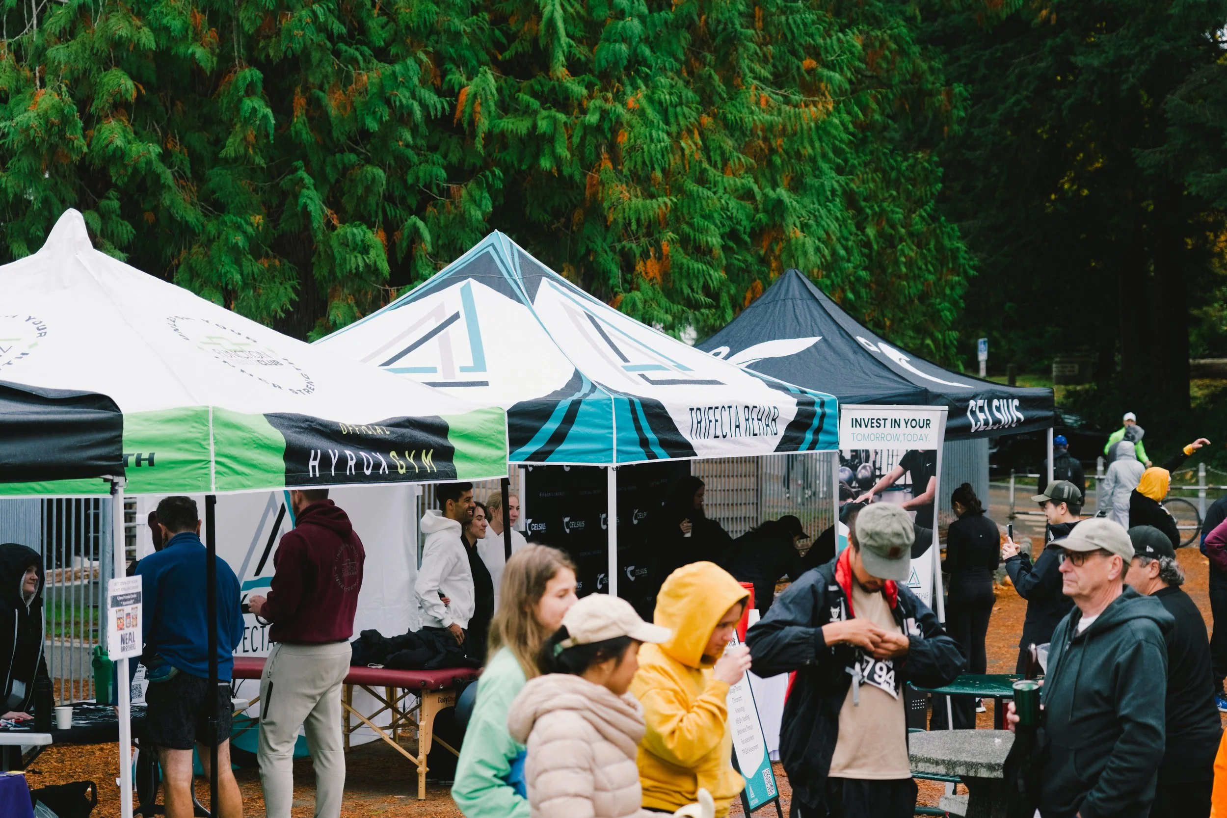 Outdoor event with multiple tents, some featuring promotional banners, and people mingling and walking in fall weather surrounded by trees.