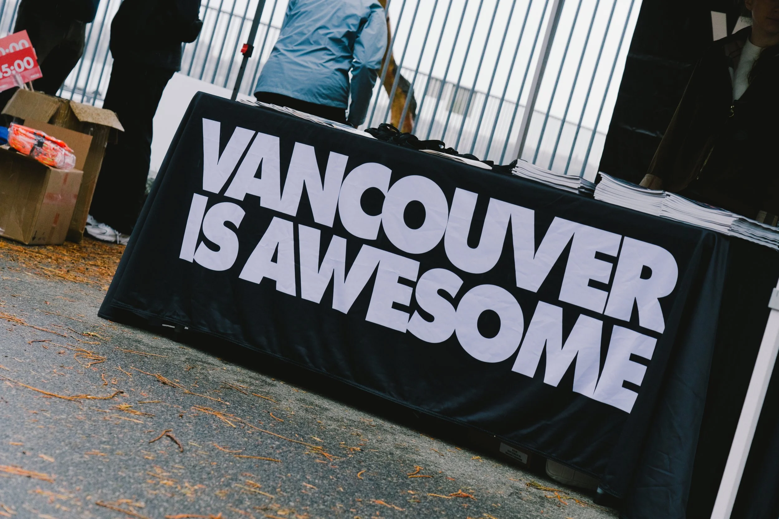 A table covered with a black cloth with large white letters that say "VANCOUVER IS AWESOME". There are stacks of papers and some boxes nearby, and people standing in the background.