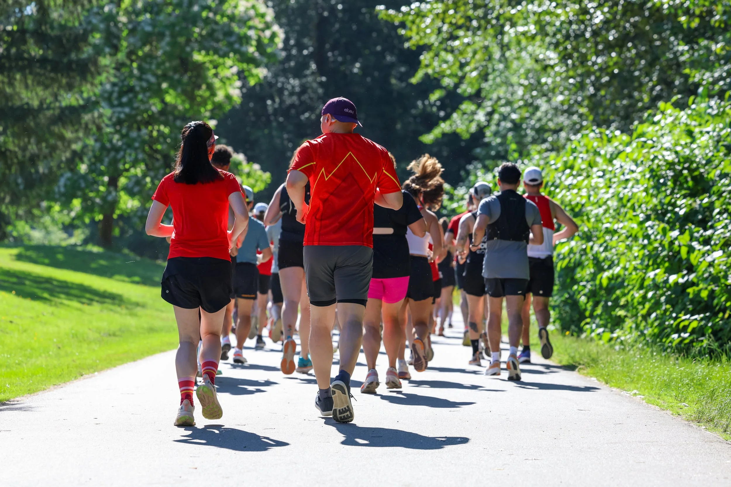 A group of people running or jogging together on a sunny outdoor trail surrounded by green trees and grass.