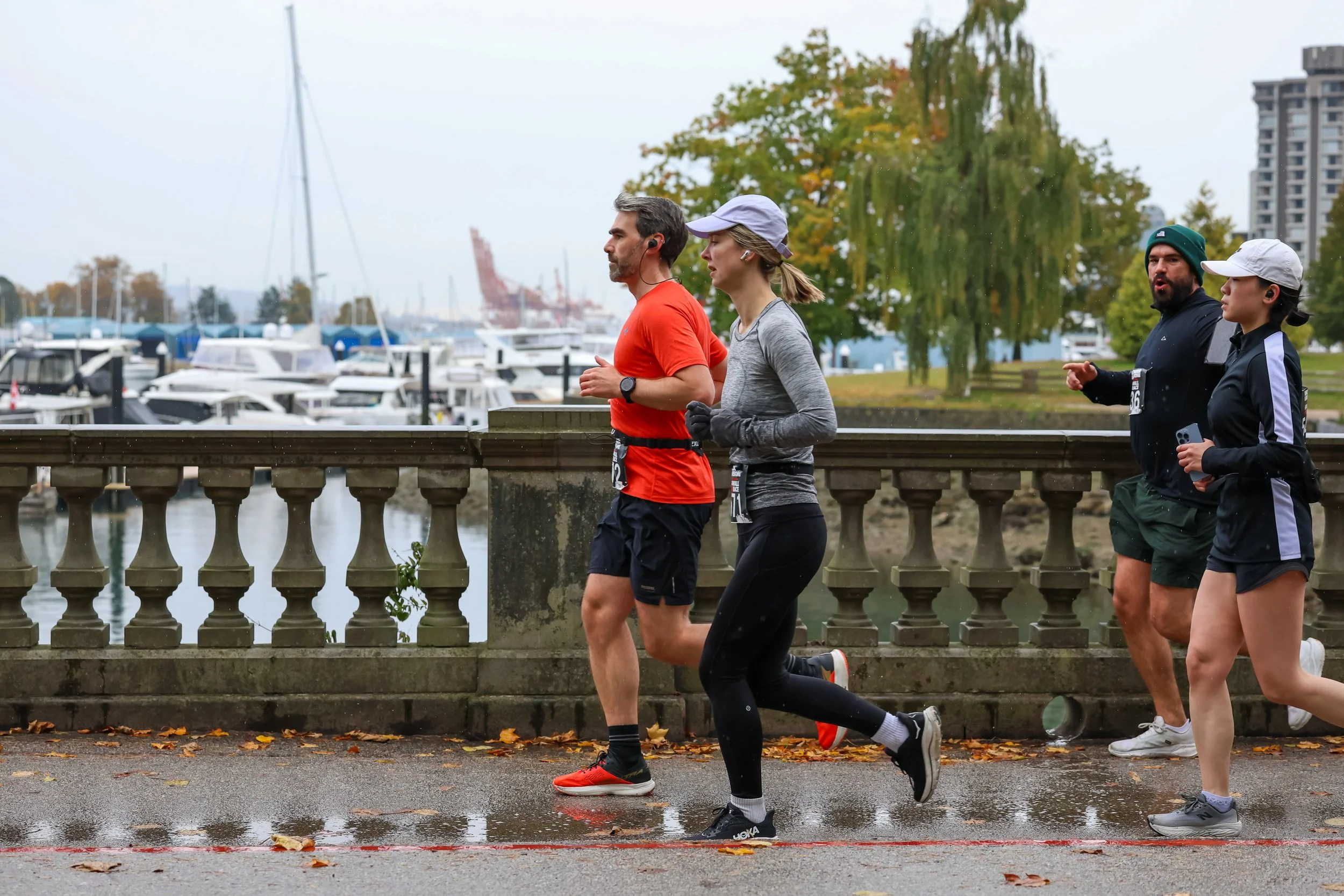 Four runners jogging along a waterfront path on a cloudy day, with boats docked in the harbor and colorful autumn trees in the background.