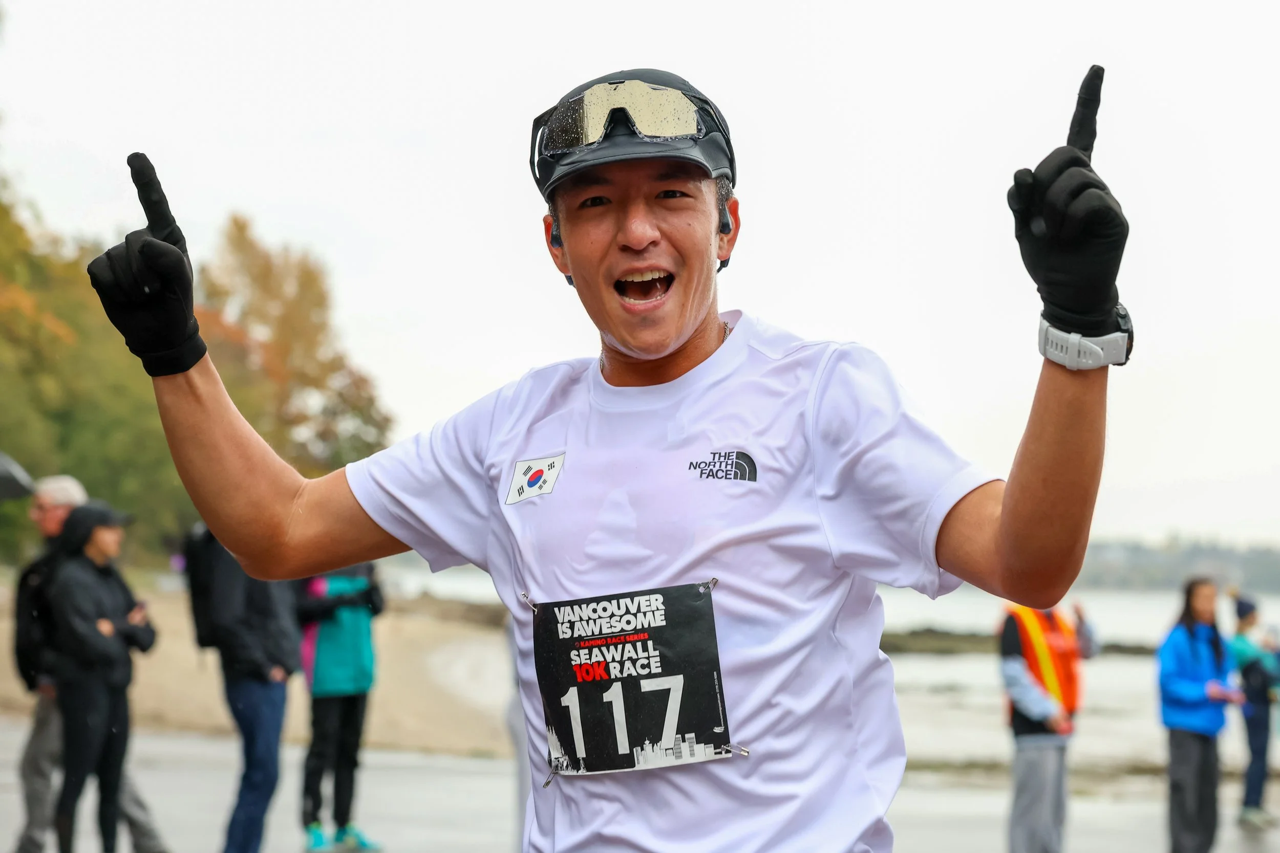 Man smiling and celebrating while running in a race, wearing a white T-shirt, black gloves, sunglasses on his head, race bib number 117, and headphones, with a scenic outdoor background and spectators in the distance.