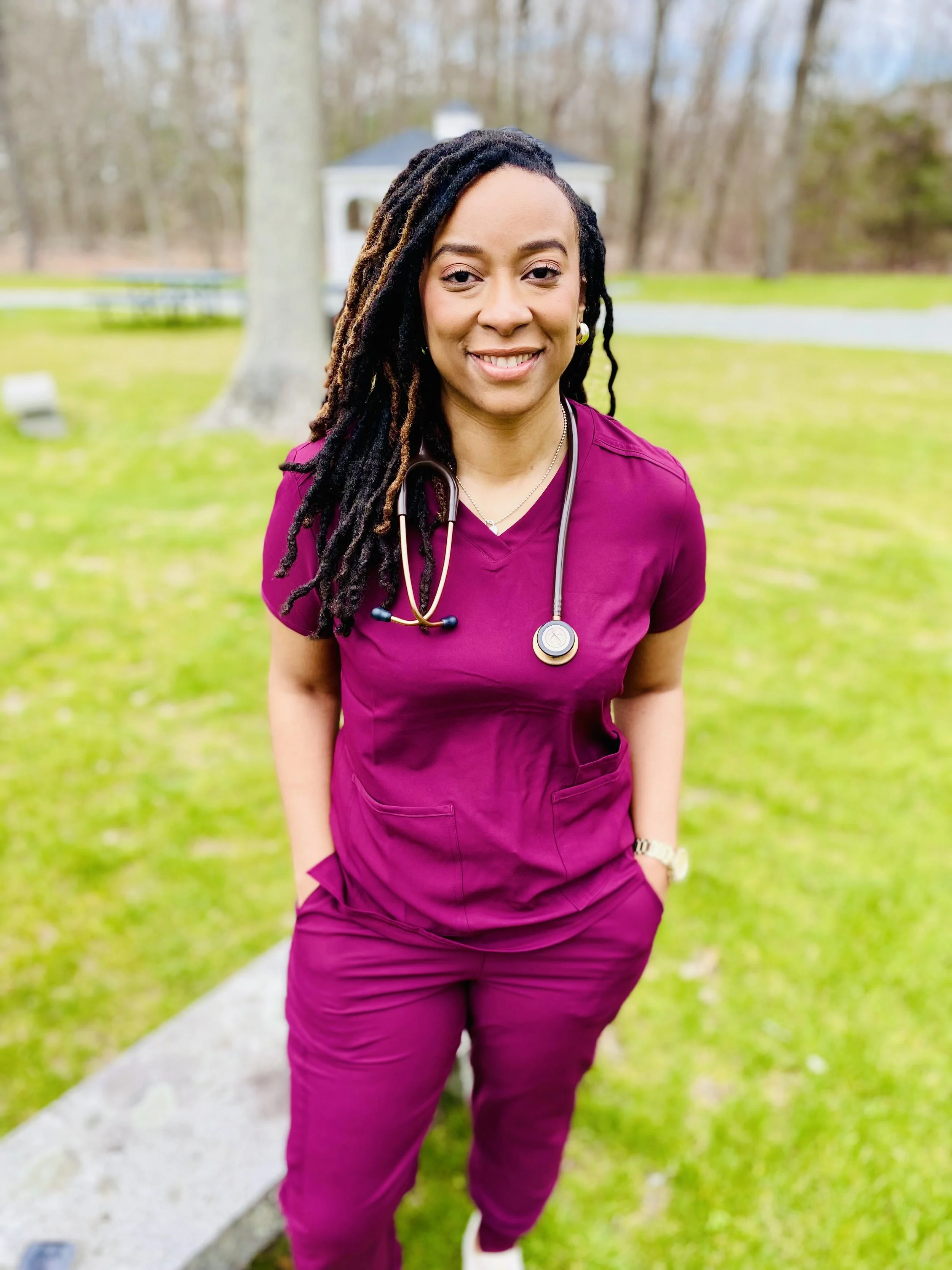 A woman in purple medical scrubs with a stethoscope around her neck standing outdoors on a grassy area with trees and a small building in the background.