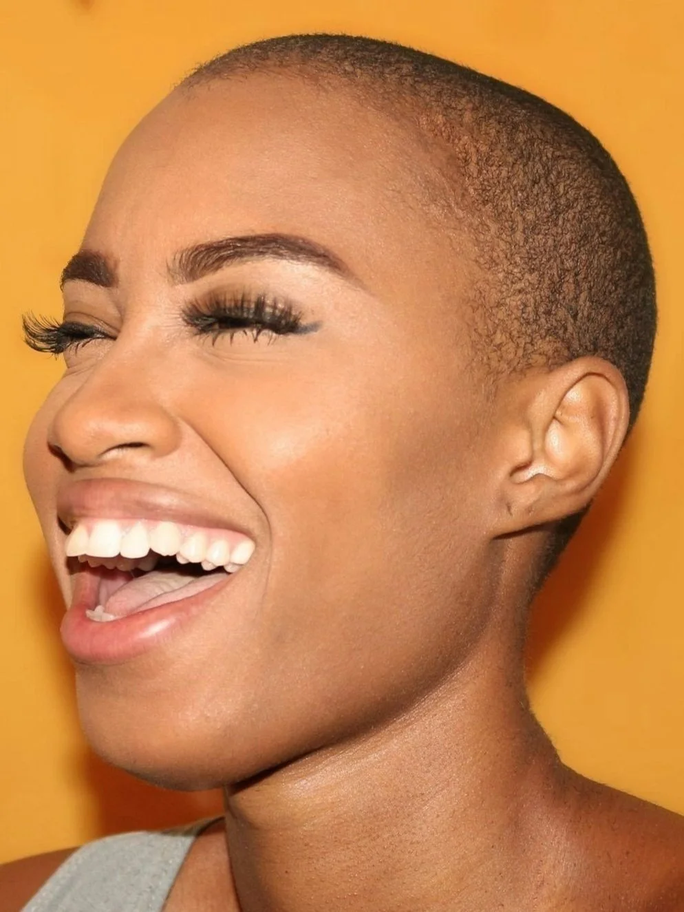 A woman with a shaved head, smiling and showing her teeth, against a yellow background.