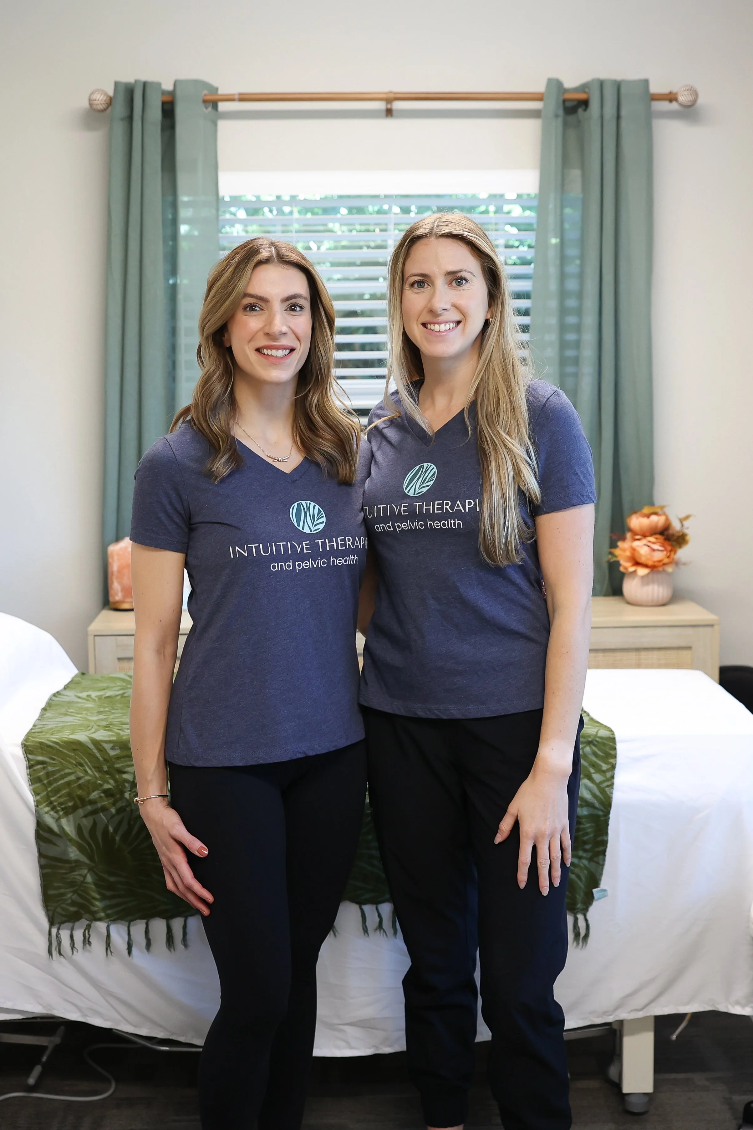 Two women in matching navy t-shirts with 'Intuitive Therapy and Pelvic Health' logo, smiling indoors with a bed and window behind them. Nicole Woodruff, OTD, OTR/L and Abby Mandelblatt, PT, DPT - the team at Intuitive Therapies & Pelvic Health