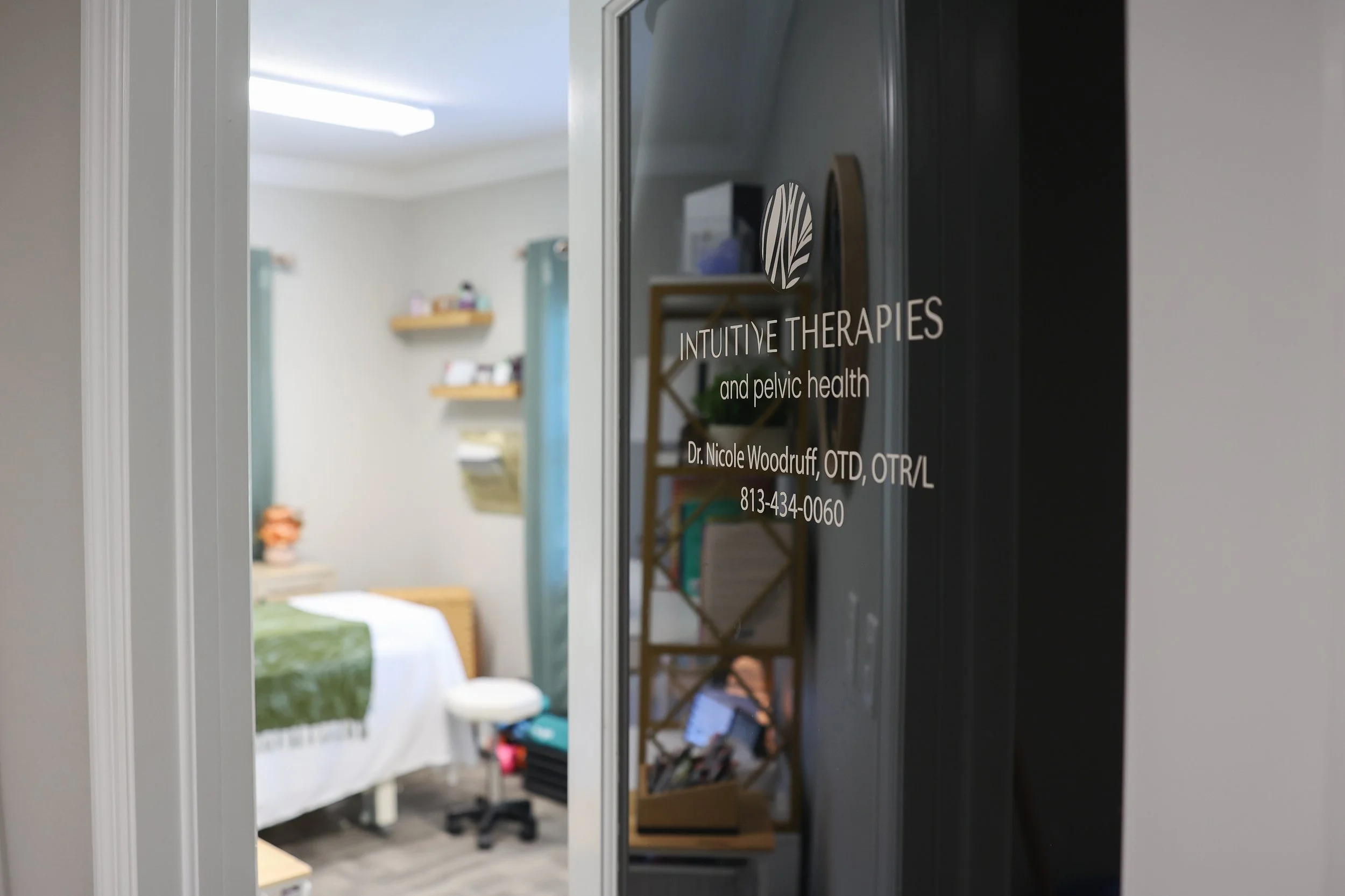 Interior of a Physical and occupational therapy treatment room with a bed, shelves, and curtains, viewed through a glass door with the Intuitive Therapies & Pelvic Health name and contact information.